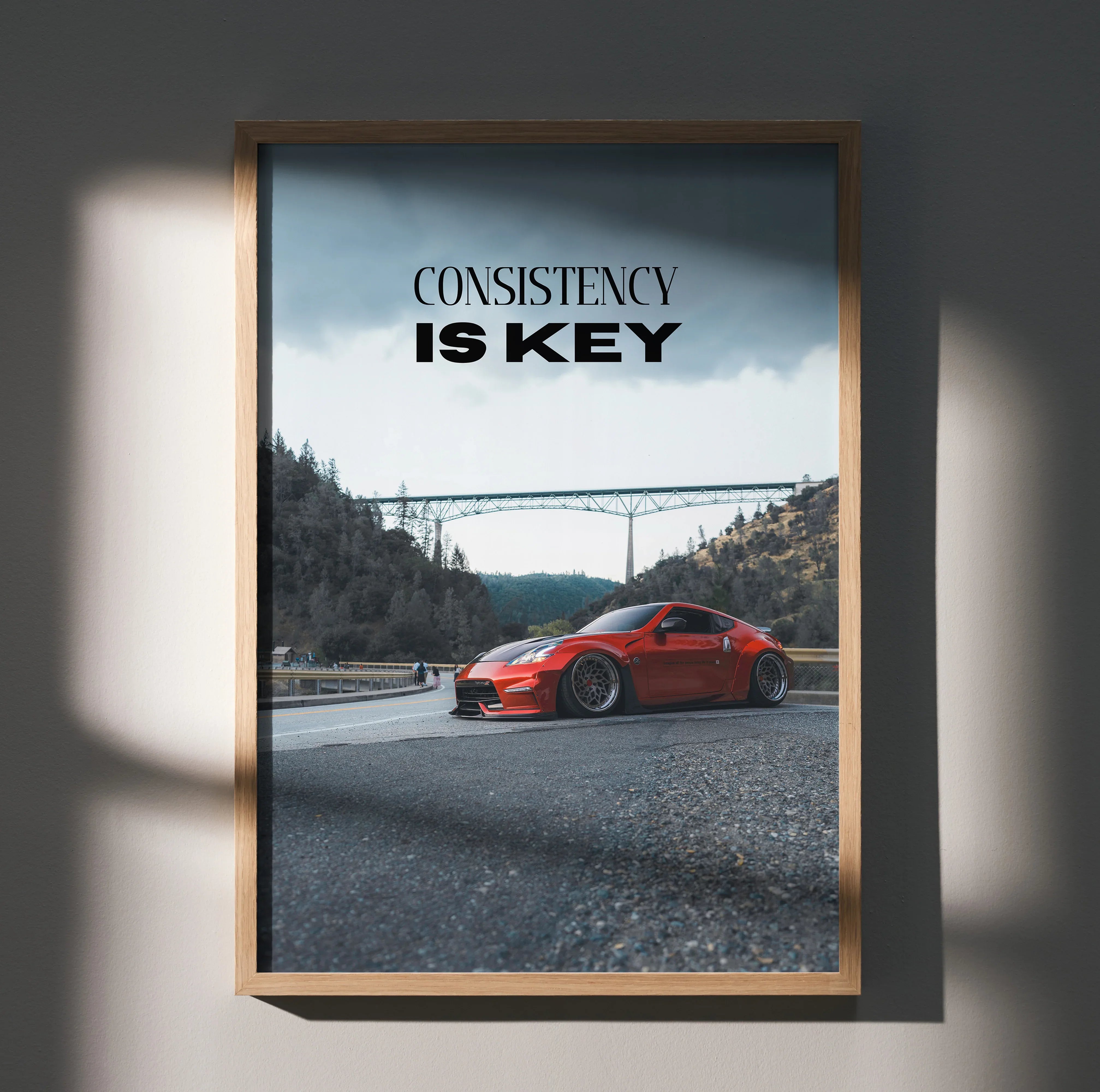 Motivational wall art poster with red Nissan 370z and text "Consistency Is Key" against mountain and bridge background.