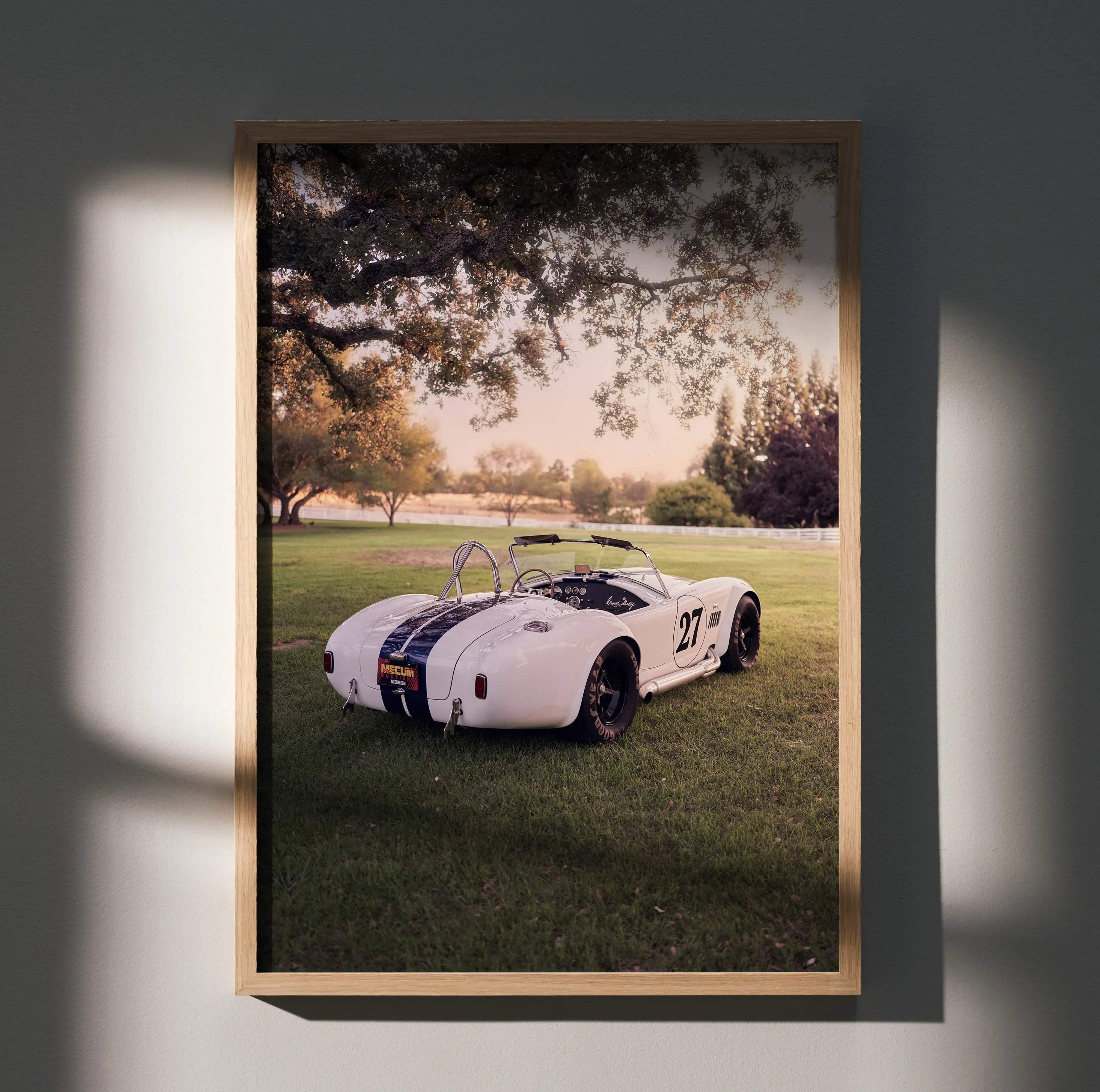 A framed photograph of a white vintage shelby cobra race car with black stripes number 27 in a light wood frame for classic car decor