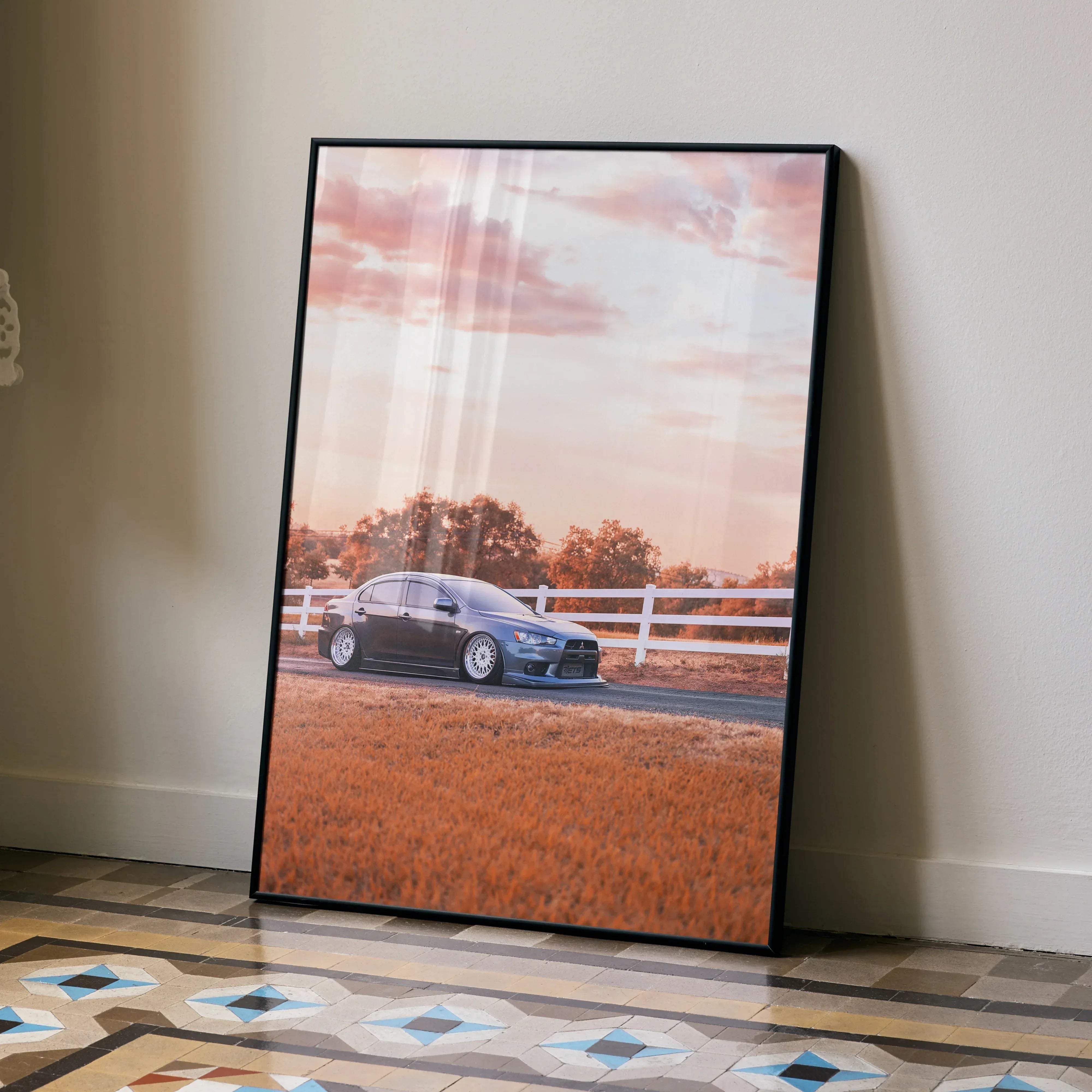 Black-framed poster featuring a dark gray mitsubishi lancer evolution parked on a rural road at sunset with white fences