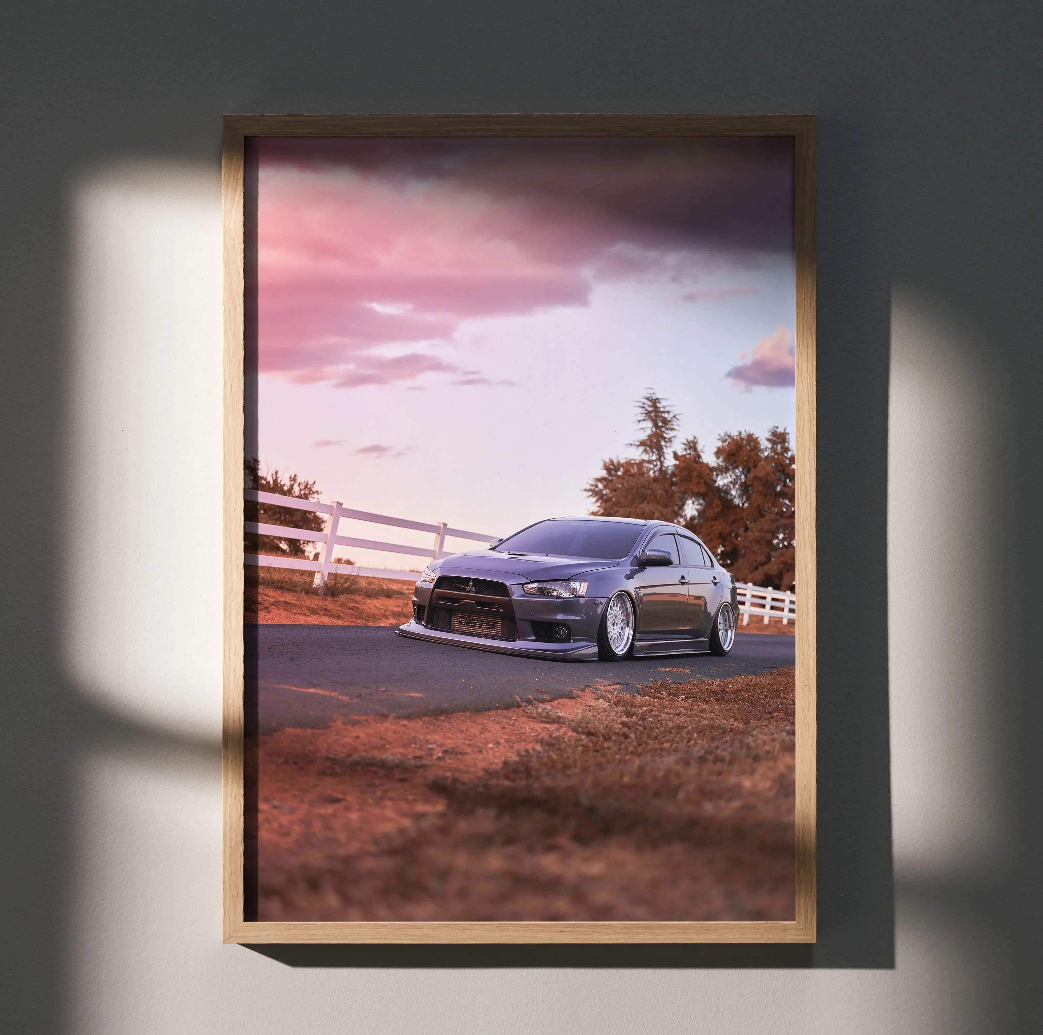 A framed poster of a dark gray Mitsubishi Lancer Evolution 10 parked on a rural road under a pink and purple sky