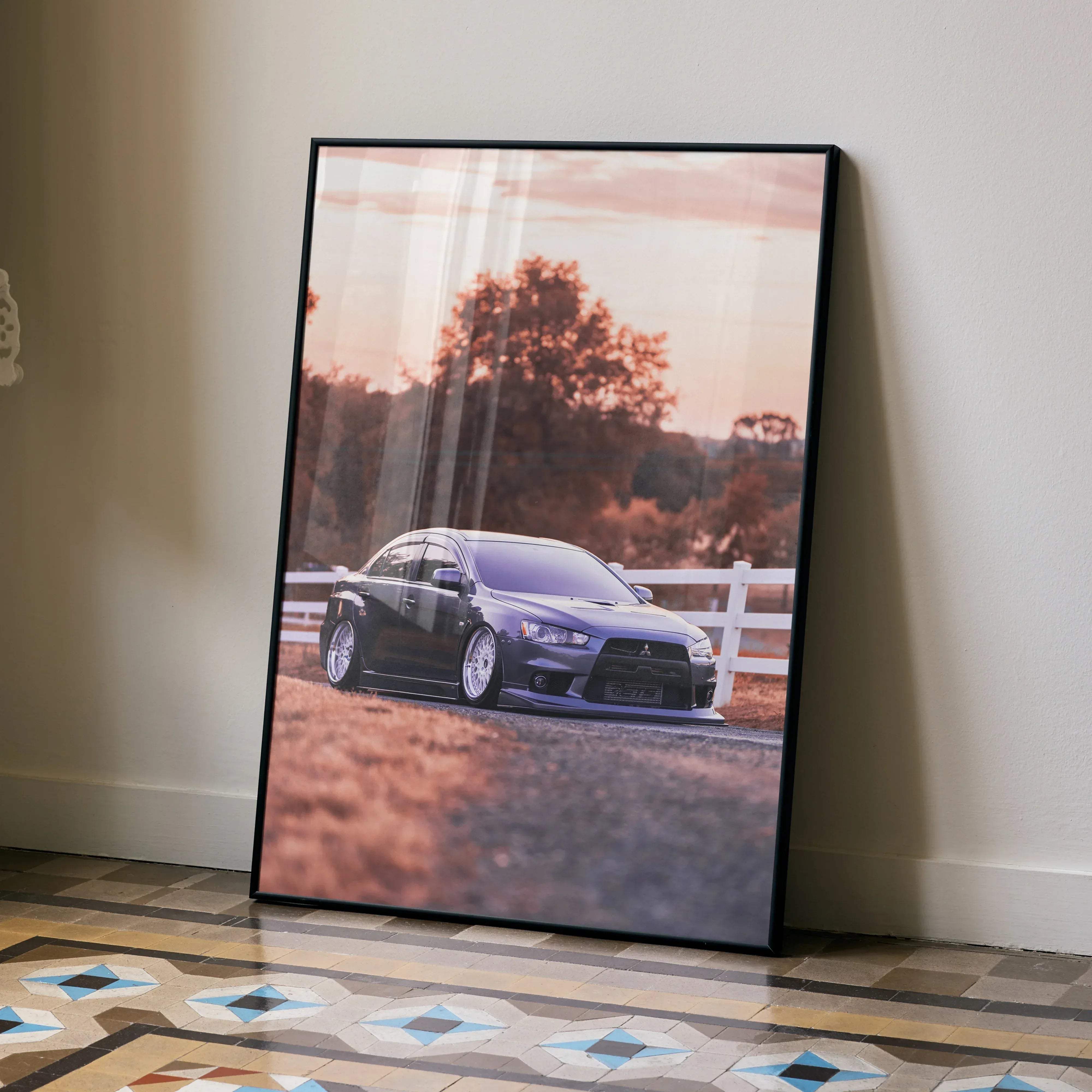 Black framed poster of a matte black mitsubishi lancer evolution parked on a dirt road with trees and white fence background