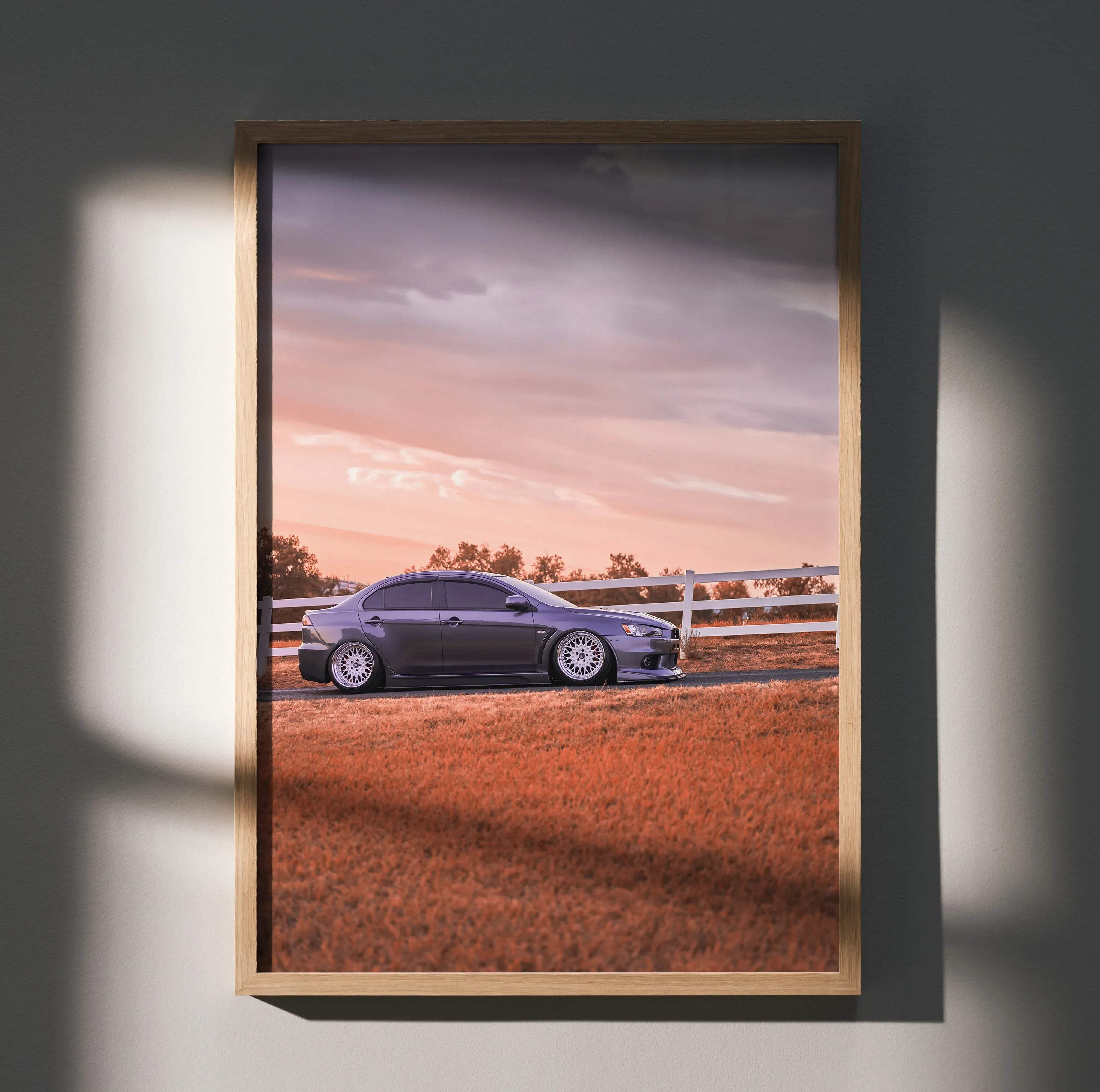 A framed high resolution poster of a dark gray Mitsubishi Lancer Evolution 10 parked by a white fence at sunset