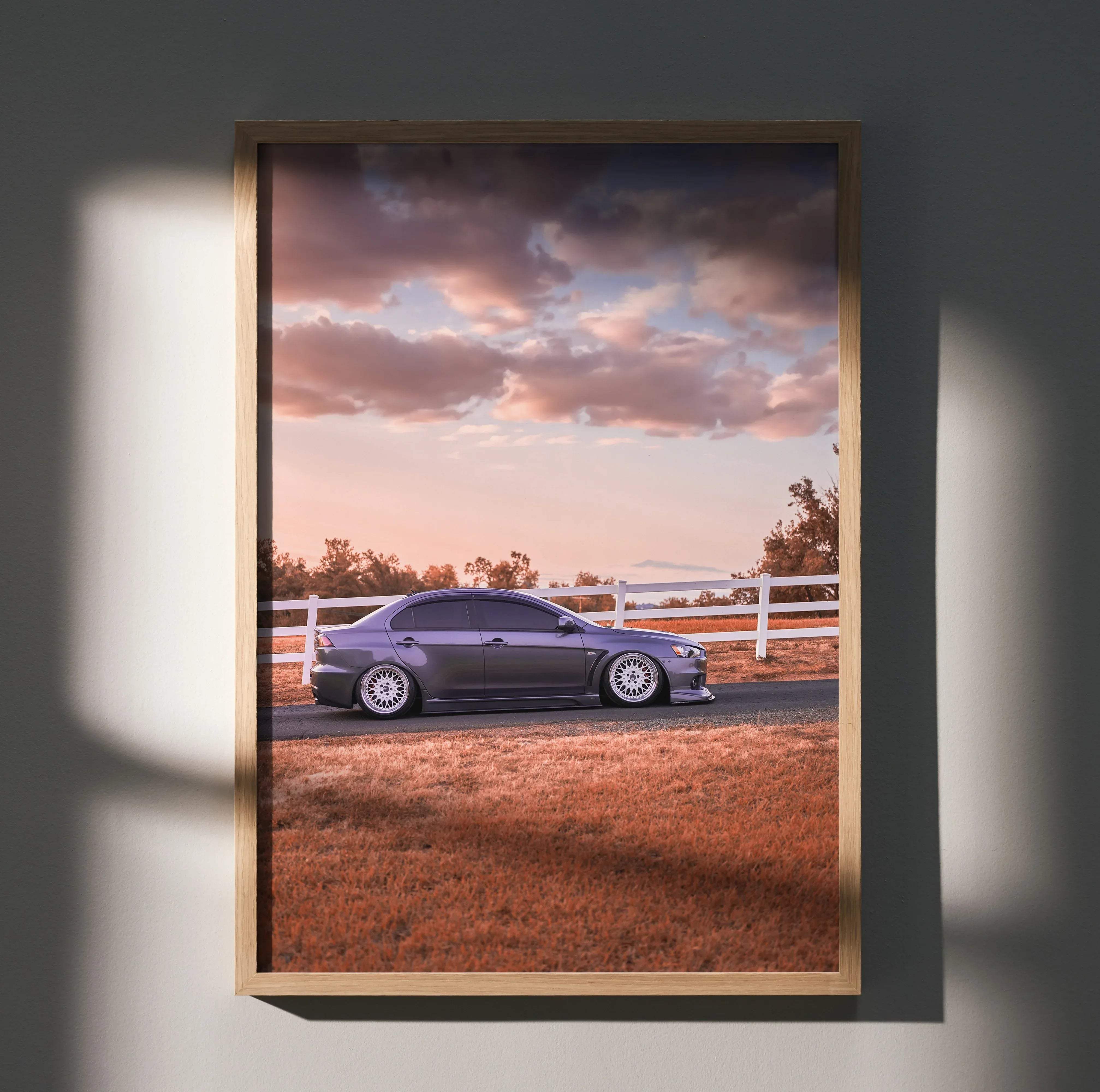A framed high resolution poster of a dark gray mitsubishi lancer evolution 10 parked beside a white fence under cloudy sky