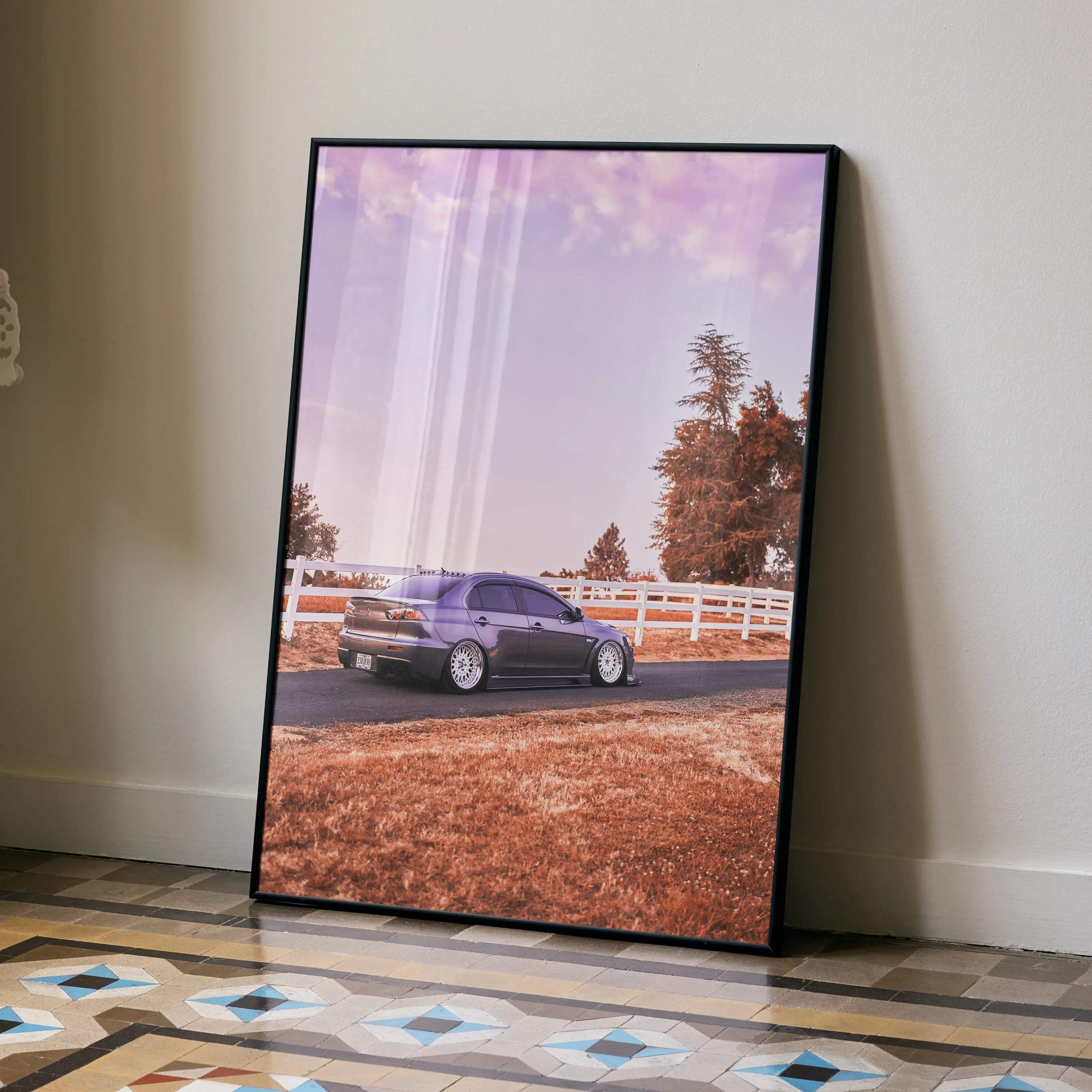 A framed photograph poster of a dark gray mitsubishi evo 10 sports car parked by trees and a white fence