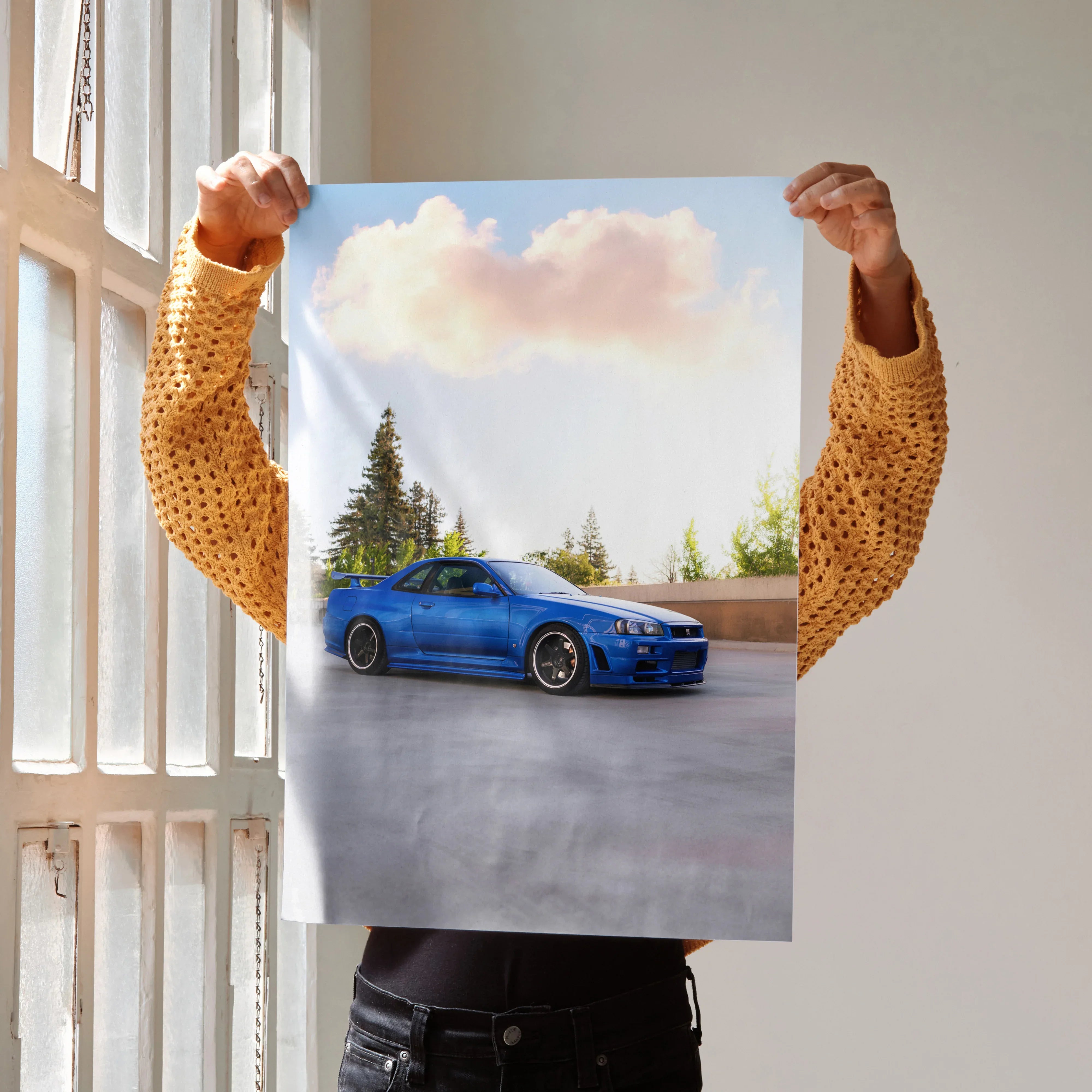 Blue Nissan R34 GTR car poster held against bright window, wall art with clear sky background.