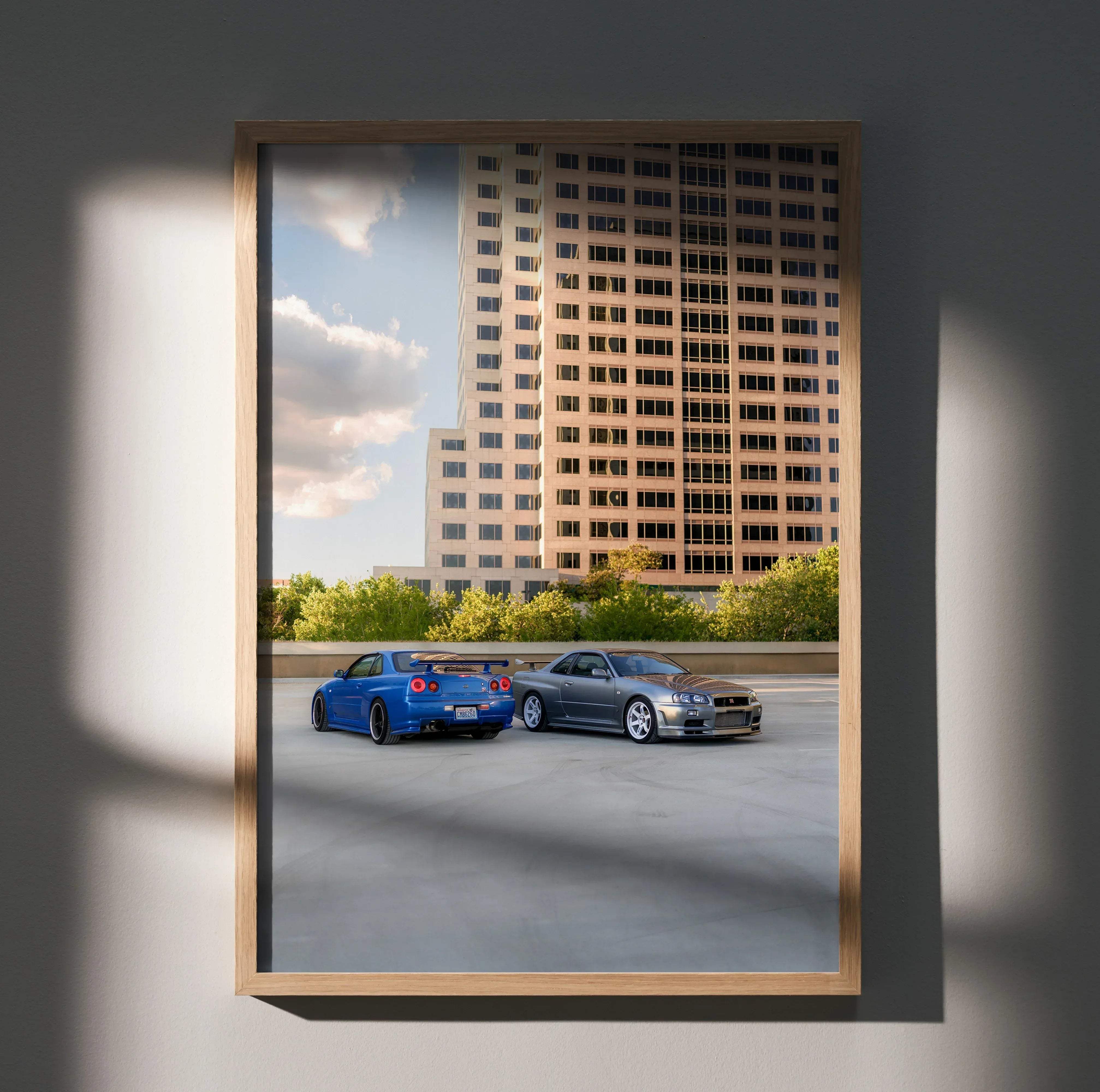 A framed Nissan R34 GTR Skyline poster featuring vibrant blue and metallic silver cars on a rooftop with city buildings