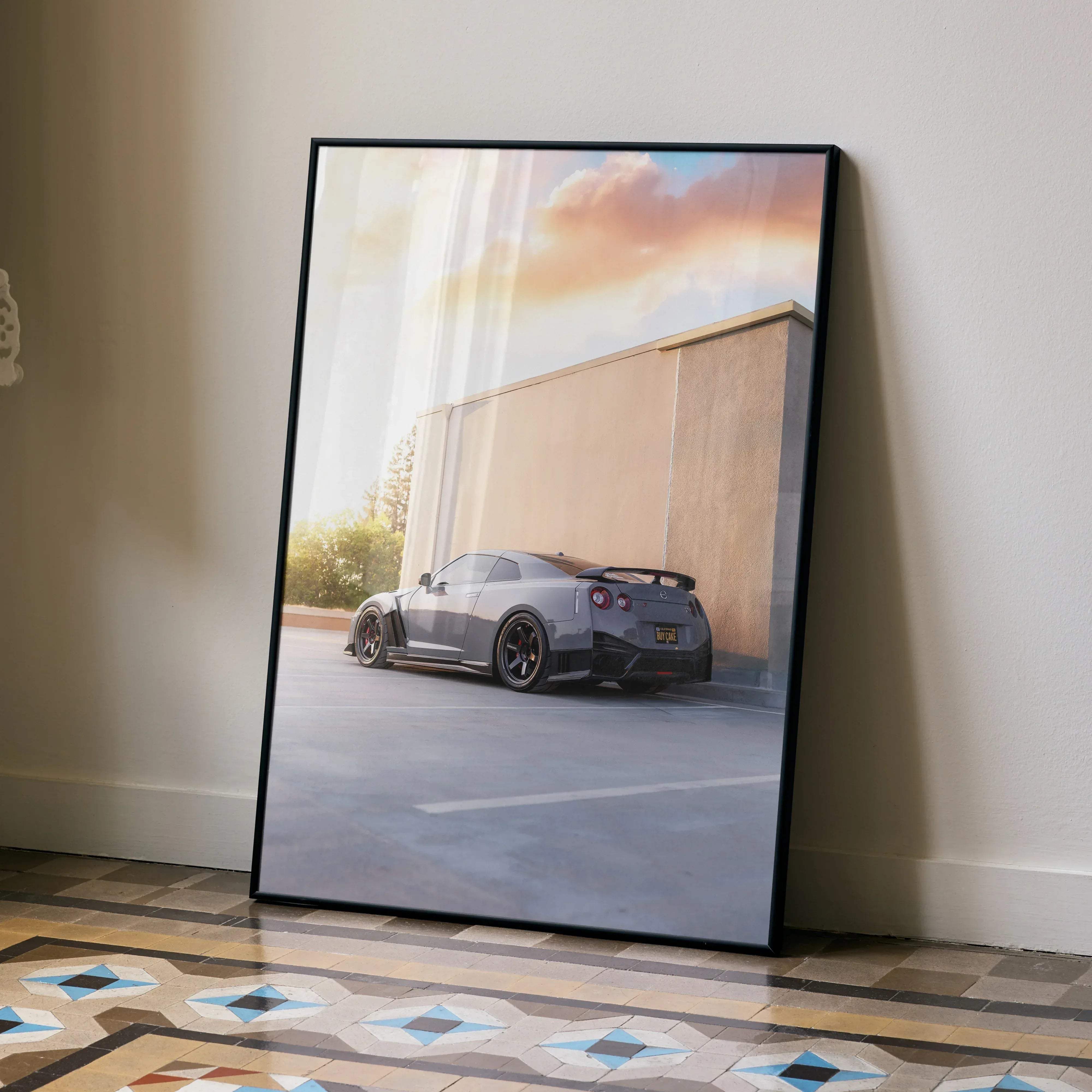 Black-framed canvas print of a gray nissan gtr r35 sports car against a beige wall under a warm cloudy sky