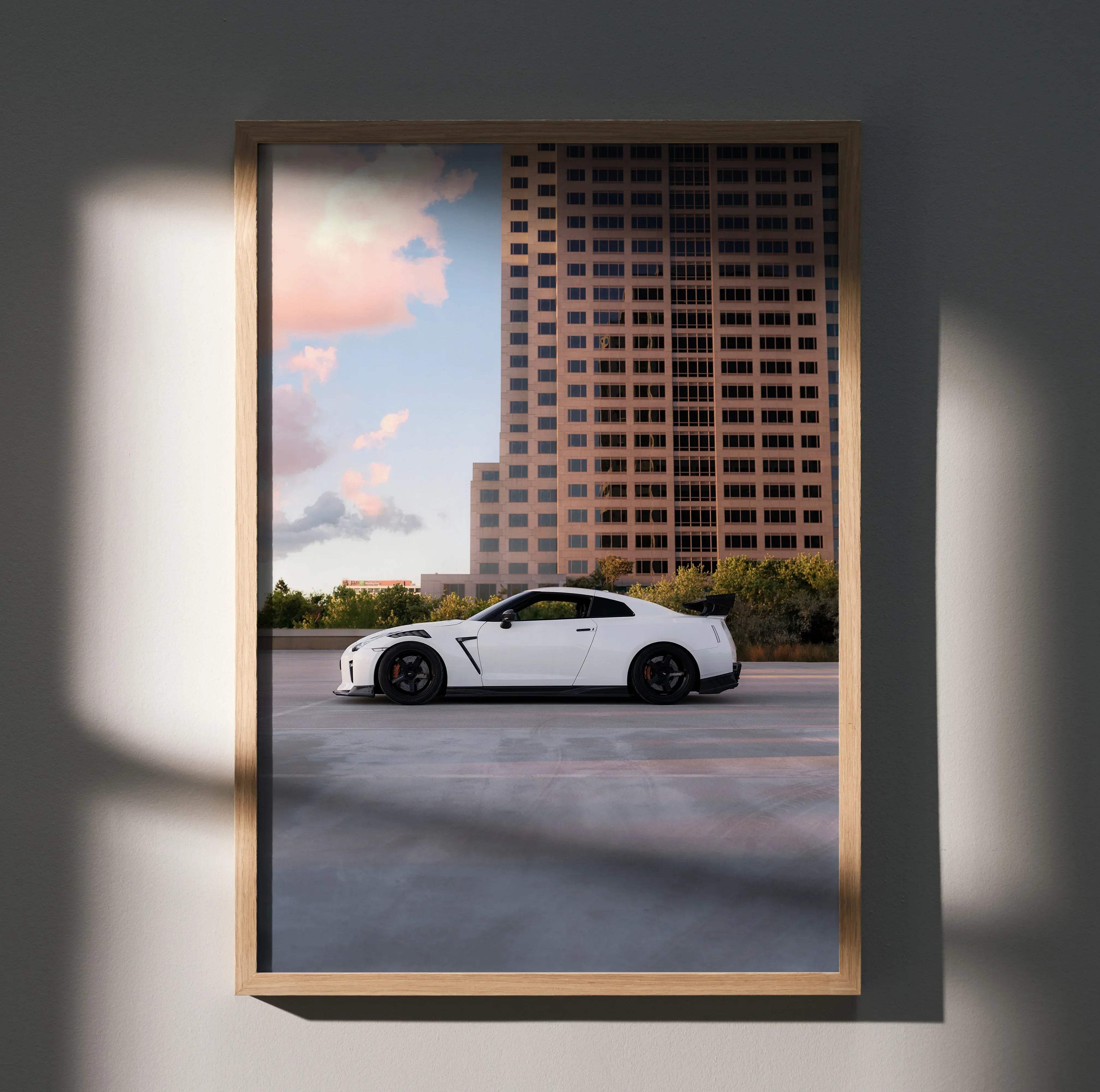 A framed poster of a white nissan gt-r r35 with black rims parked by a tall building under a pink and blue sky