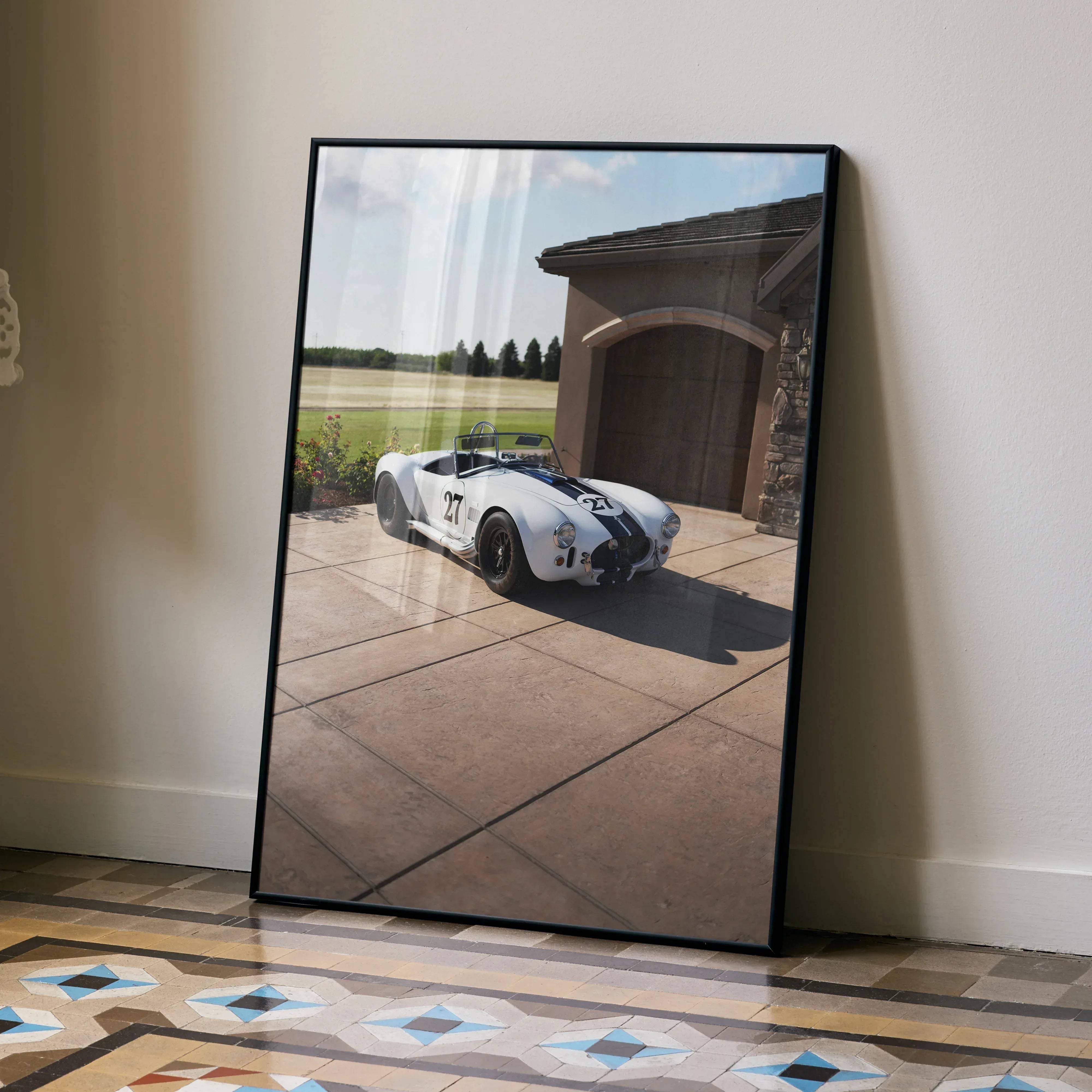 Black-framed print of a white and black number 27 racing car parked in front of a stone garage shelby cobra poster