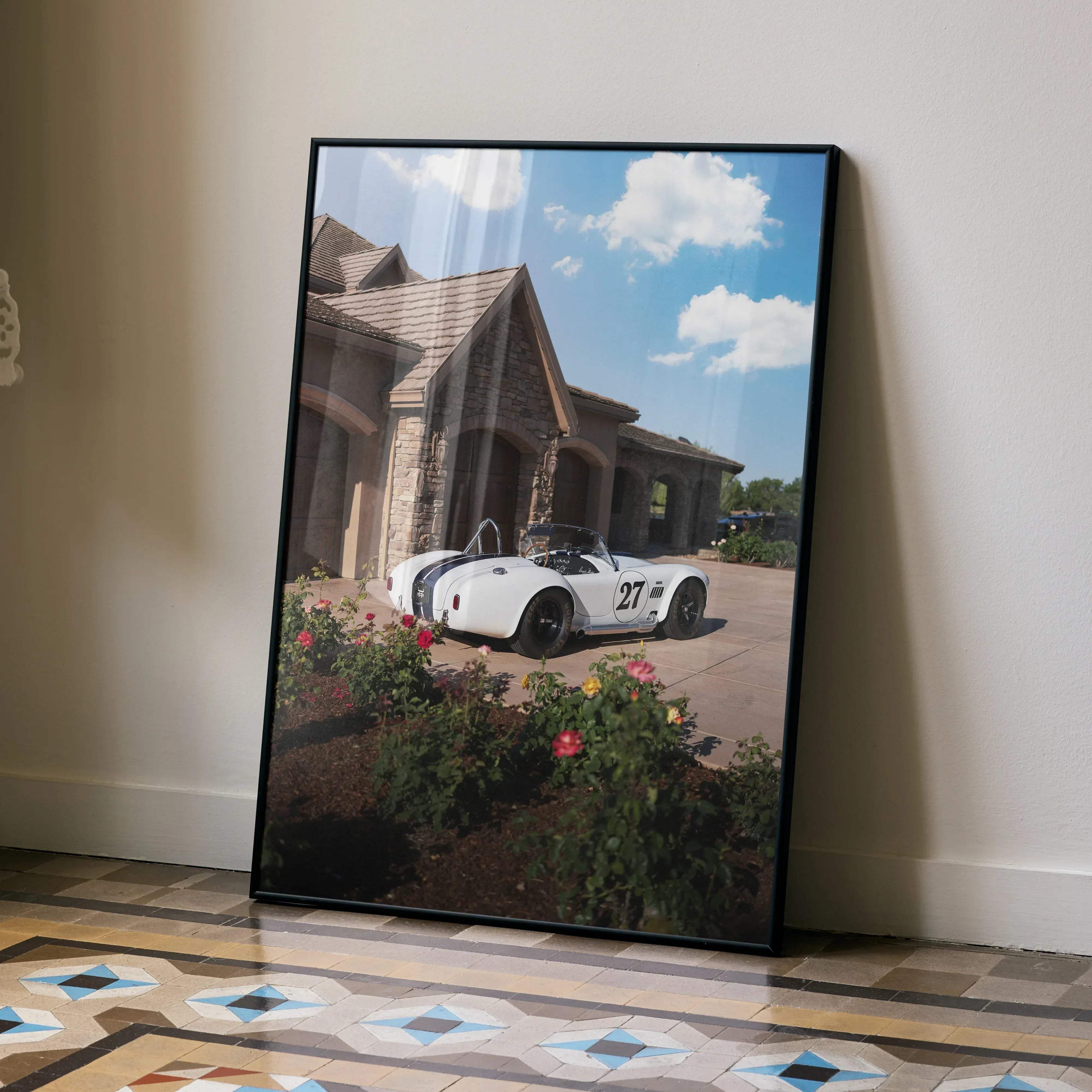 Black-framed print of a white vintage shelby cobra race car number 27 parked by a stone house under blue sky