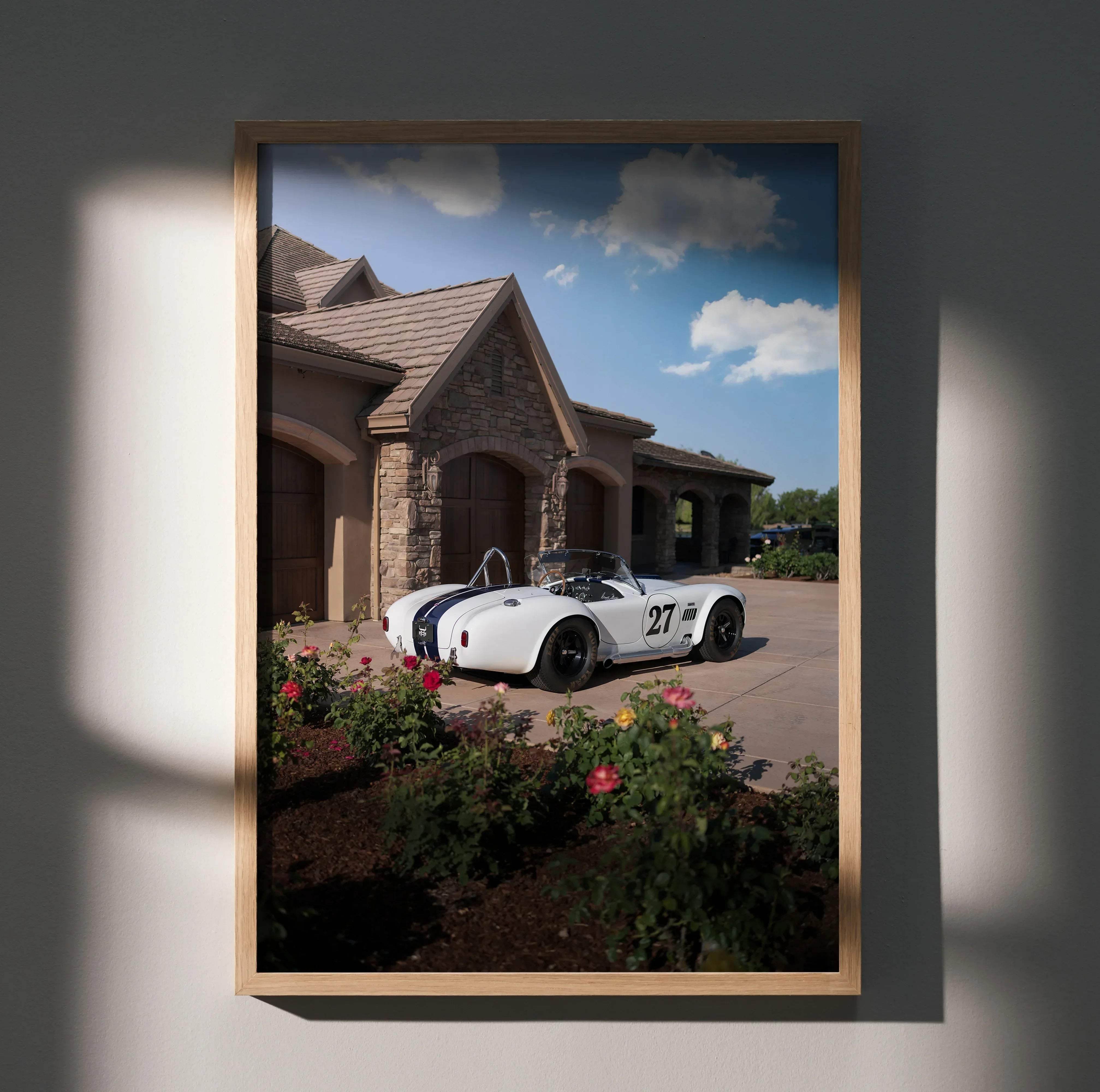 A framed high resolution car poster of a white shelby cobra with blue stripes and number 27 in front of a stone house