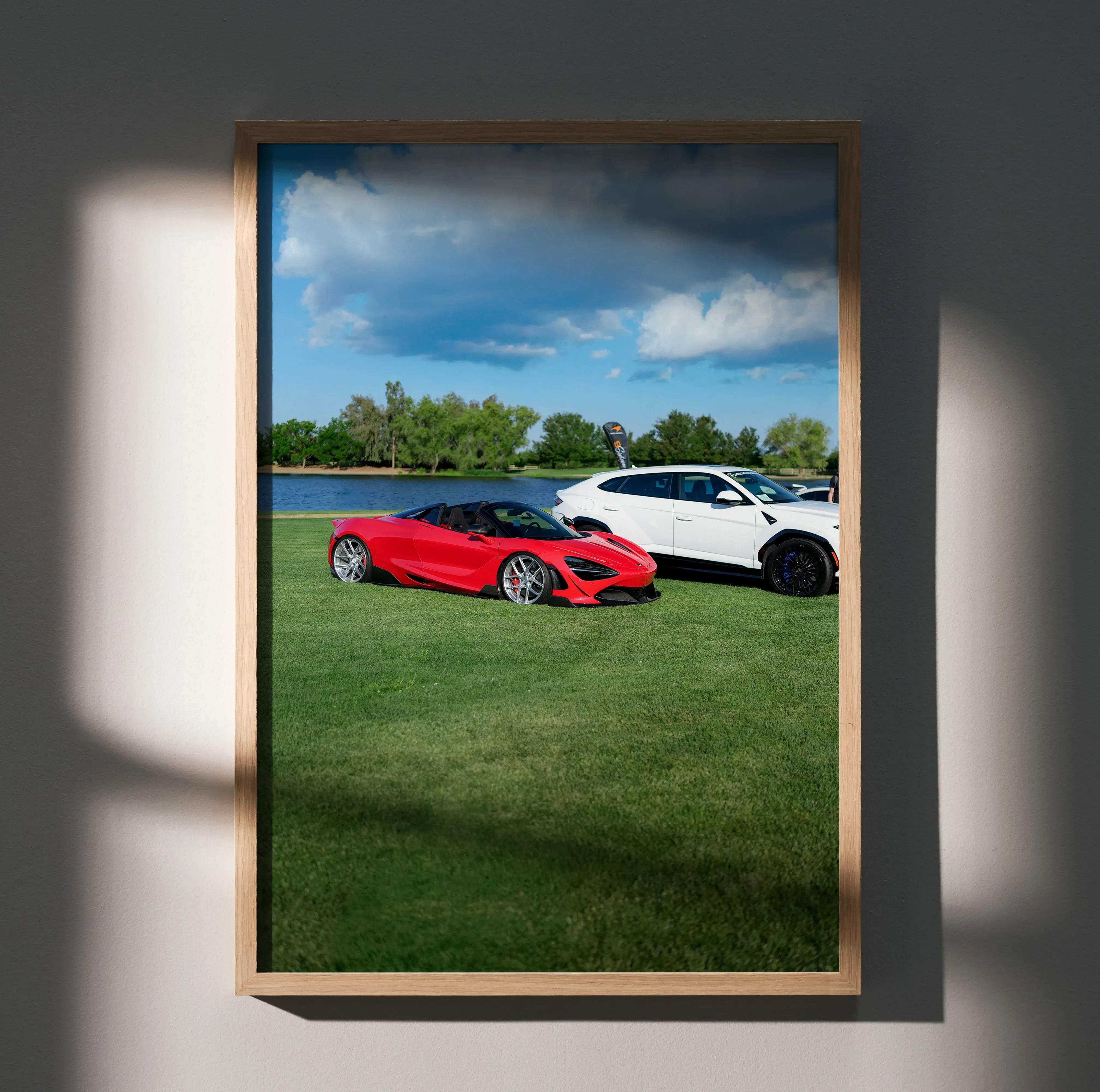 A framed photograph poster of a vibrant red McLaren 720S sports car parked on grass beside a white SUV