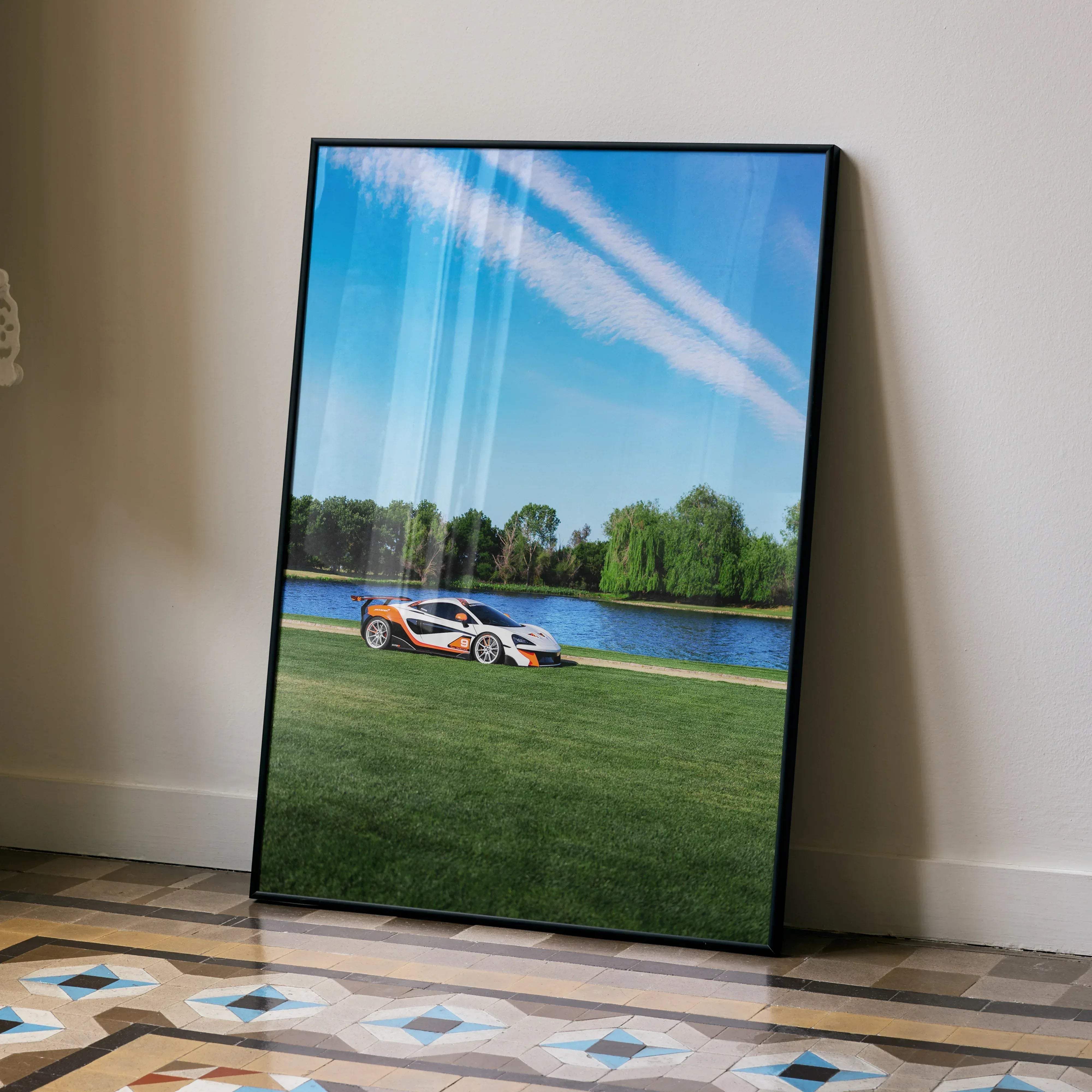 Black-framed canvas print of white and orange mclaren 570s sports car parked on grass by lake under blue sky with contrails