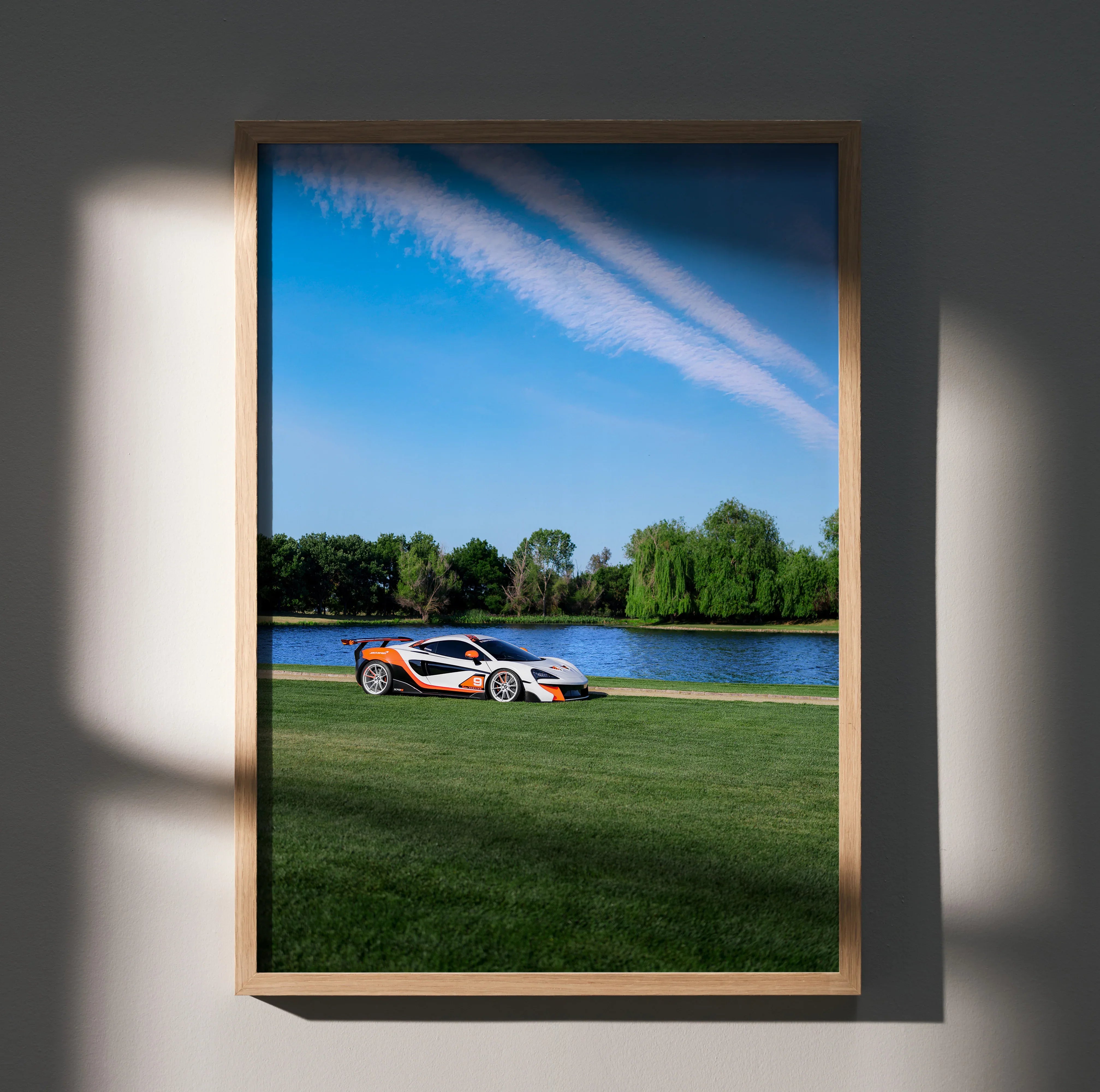 McLaren 570S poster wall art featuring the car on grass by a lake under a clear blue sky.