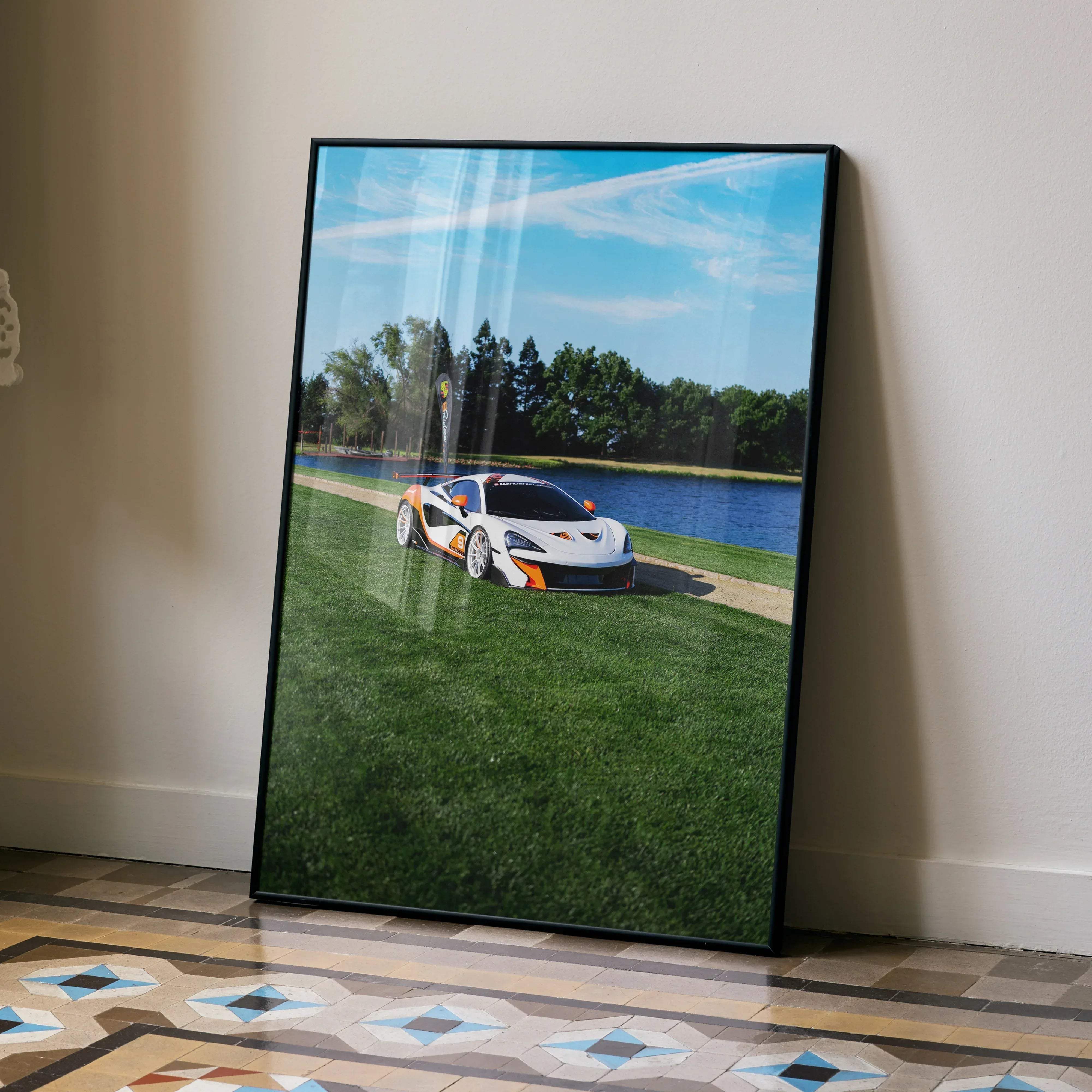 A framed poster of a white and orange mclaren 570s sports car parked on grass beside water under a blue sky