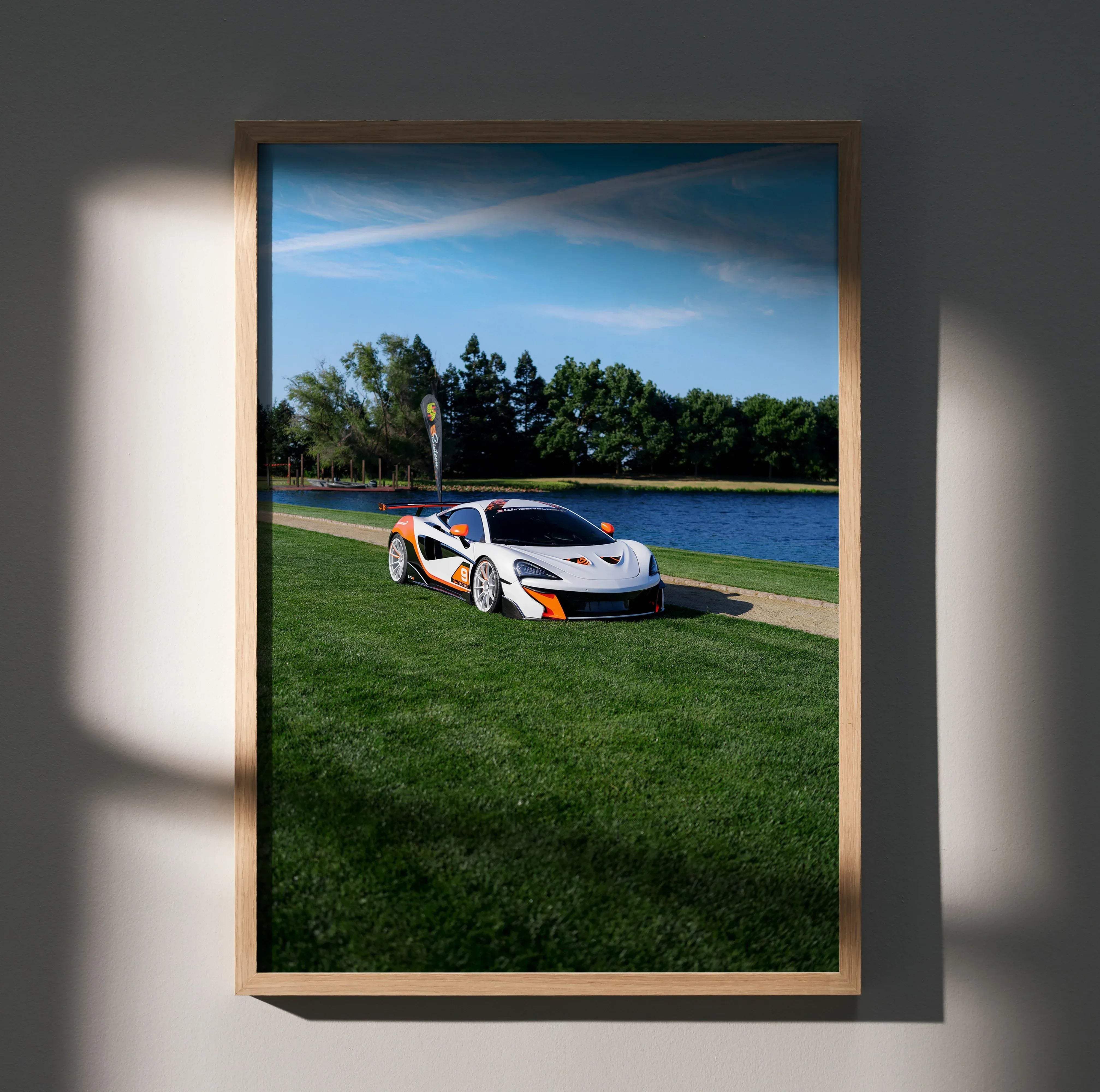 A framed photograph of a white and orange McLaren 570S sports car parked on grass beside water in high resolution