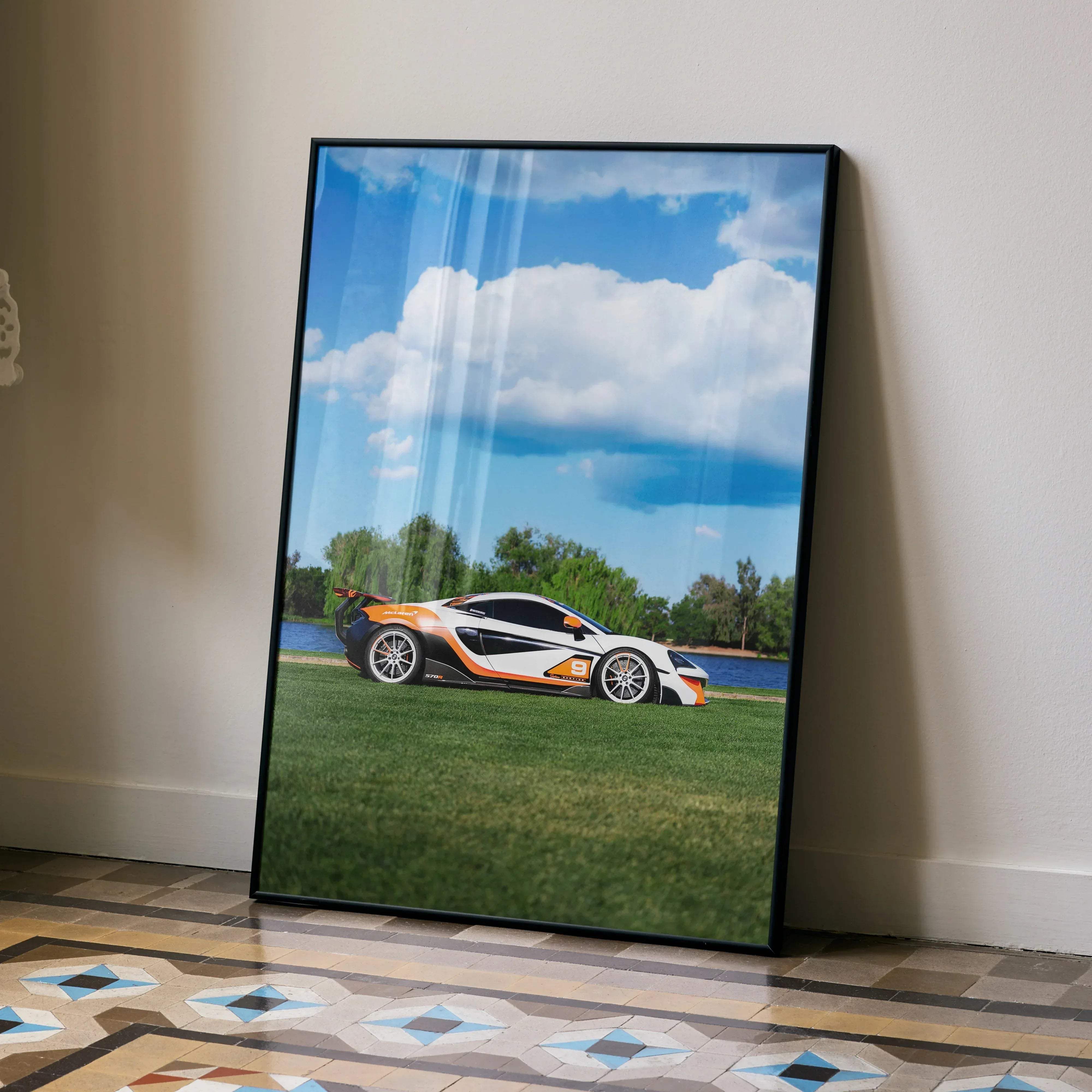 A framed poster of a white and orange mclaren 570s sports car with black accents against grass and blue sky background