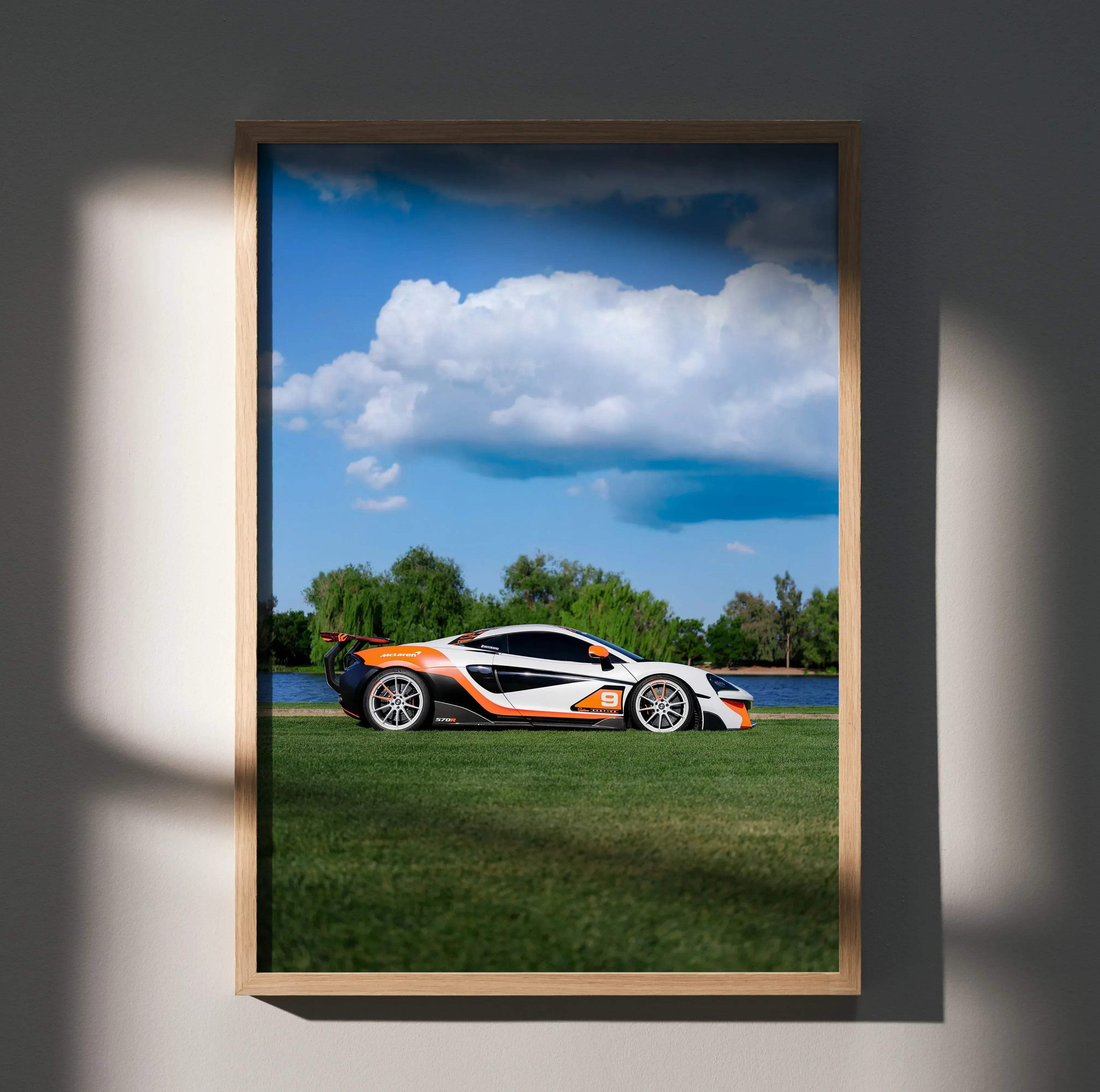 A framed photograph of a white and orange mclaren 570s sports car parked on grass under a blue sky with clouds