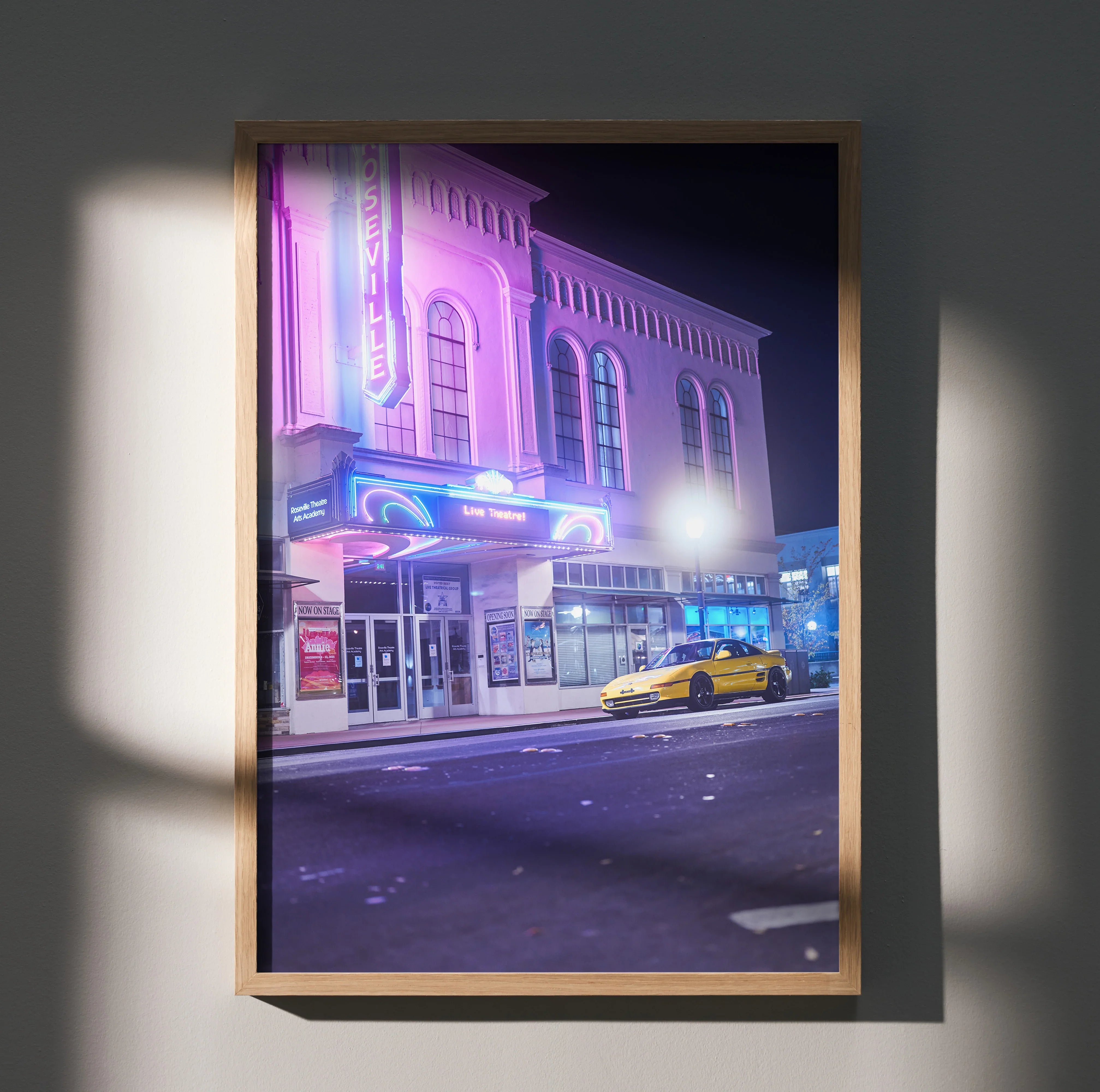 Toyota MR2 yellow sports car parked by neon-lit theater poster wall art in nighttime city.