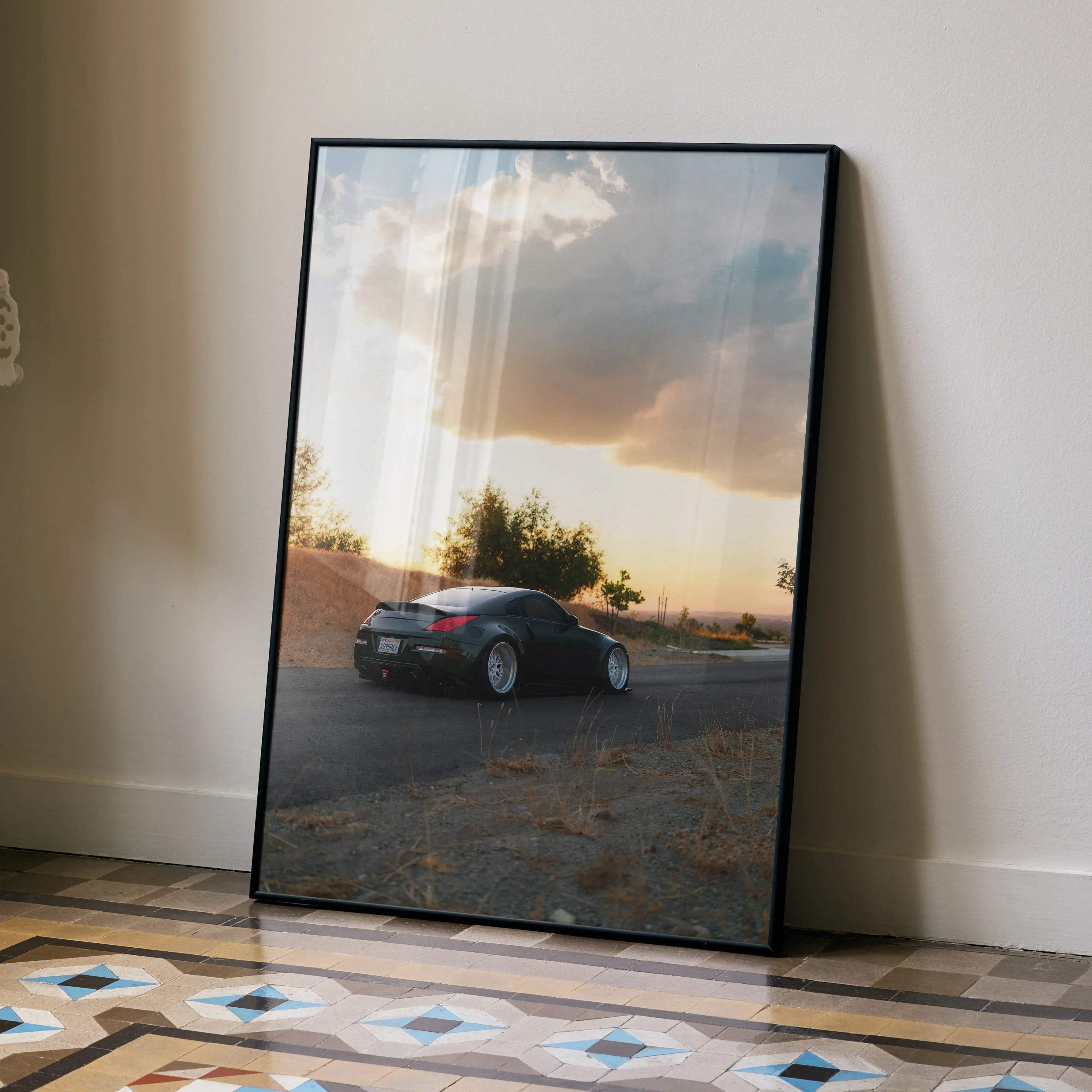 Black-framed poster featuring a sleek black nissan 350z sports car on a road at sunset with dramatic clouds and warm lighting