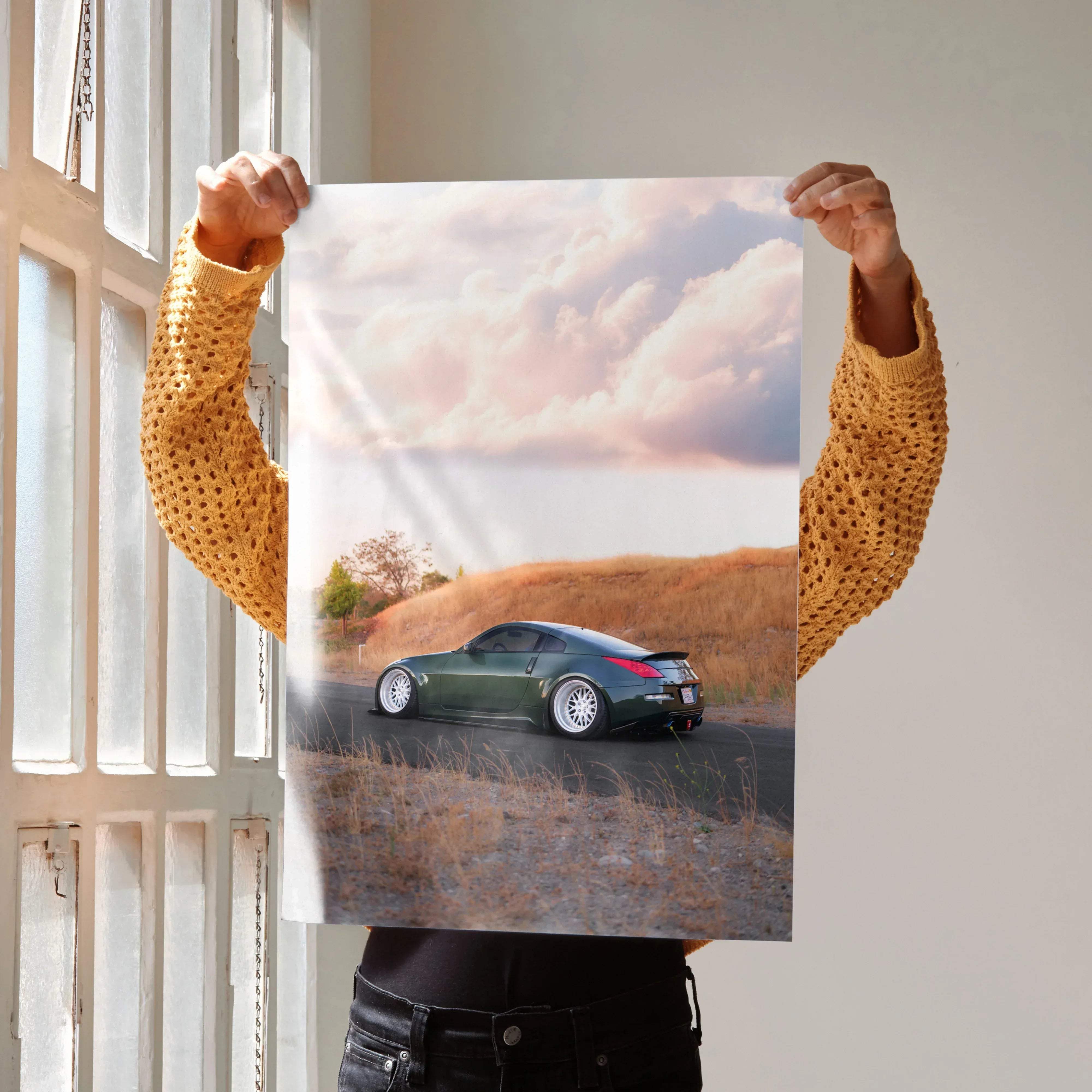 A matte-finish poster of a dark green Nissan 350z sports car with white rims on a sunlit road under pink sky