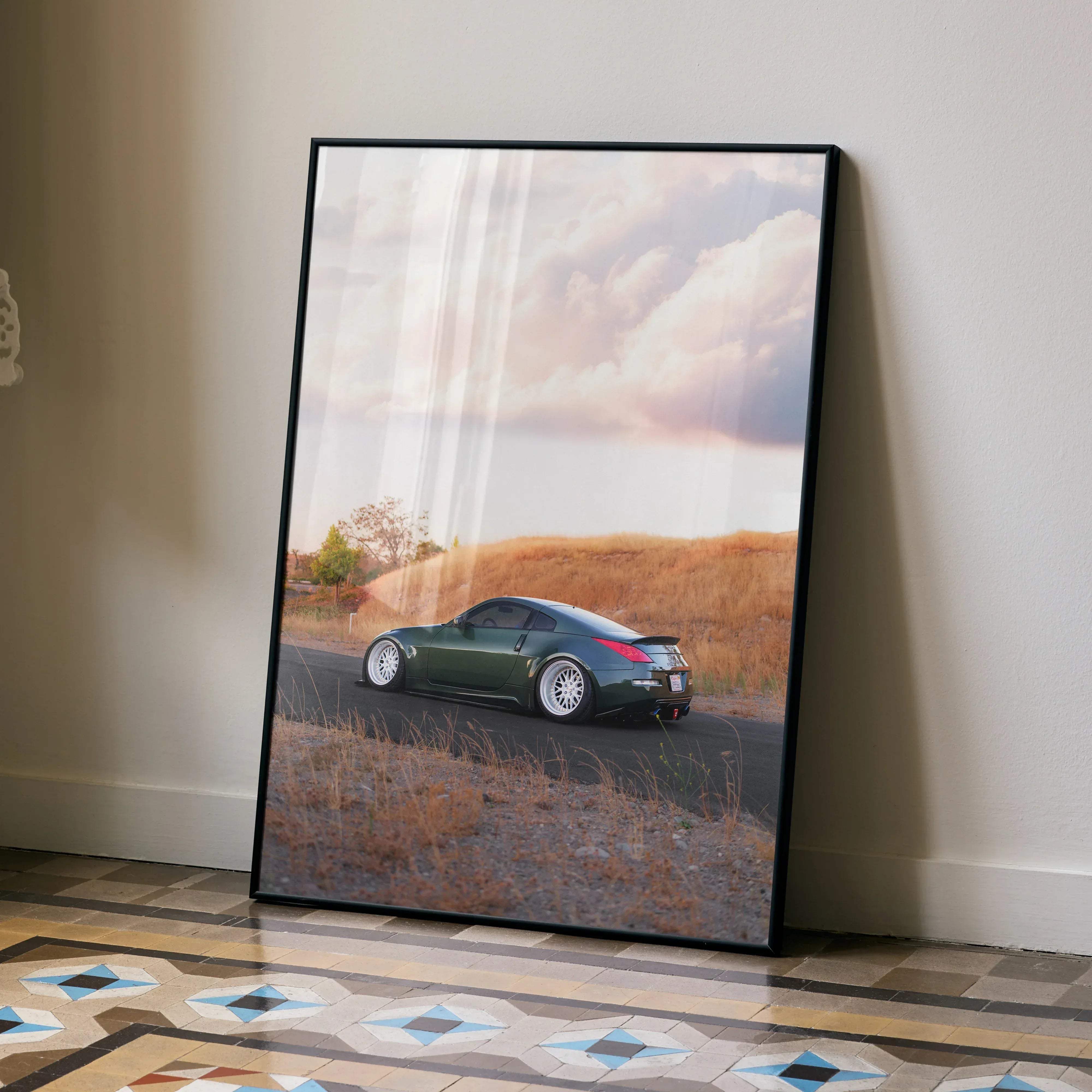 Black-framed poster of a green nissan 350z sports car with white rims on a road with golden grass under cloudy sky