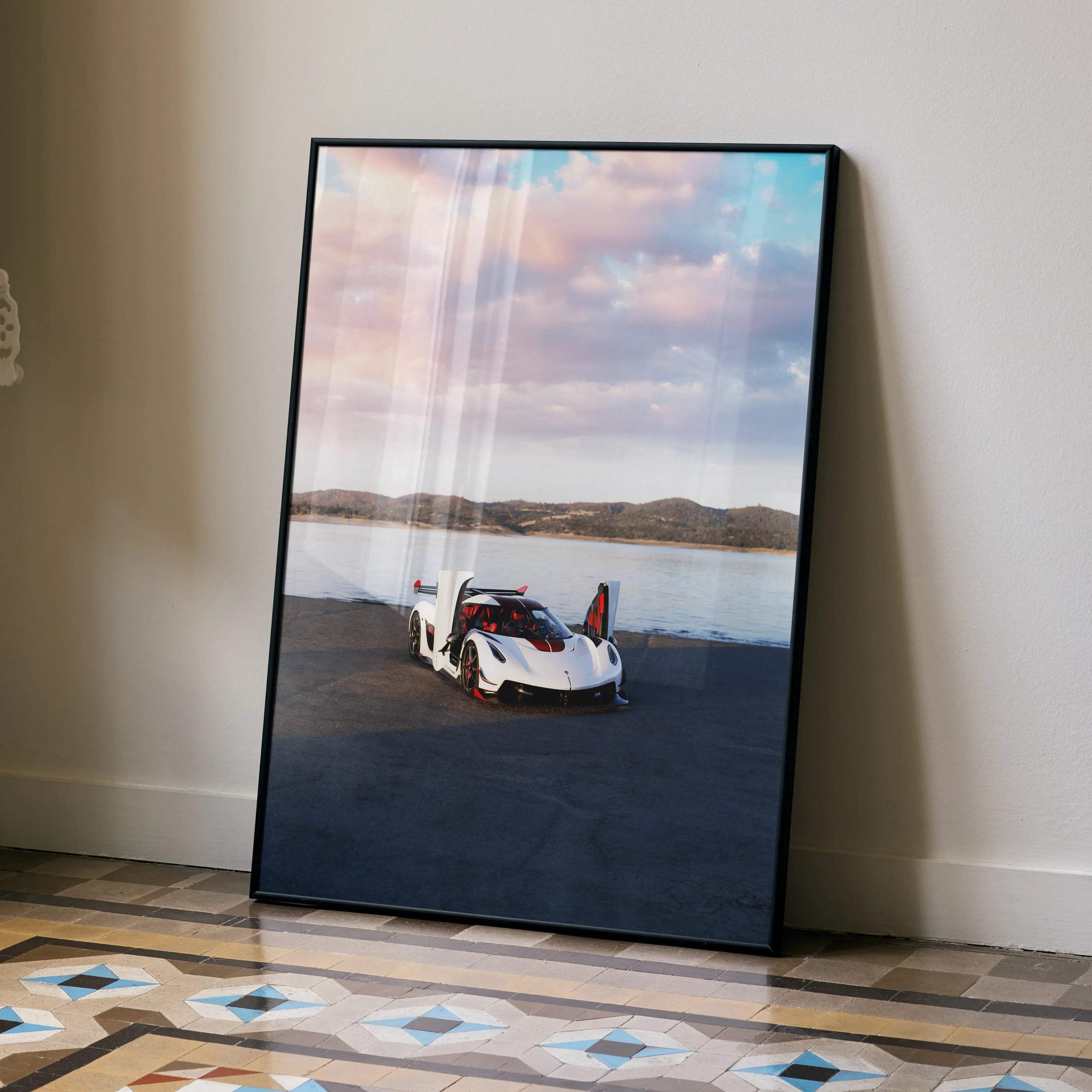 A framed poster of a white and red koenigsegg jesko hypercar with open doors by a lake under a pink and blue sky