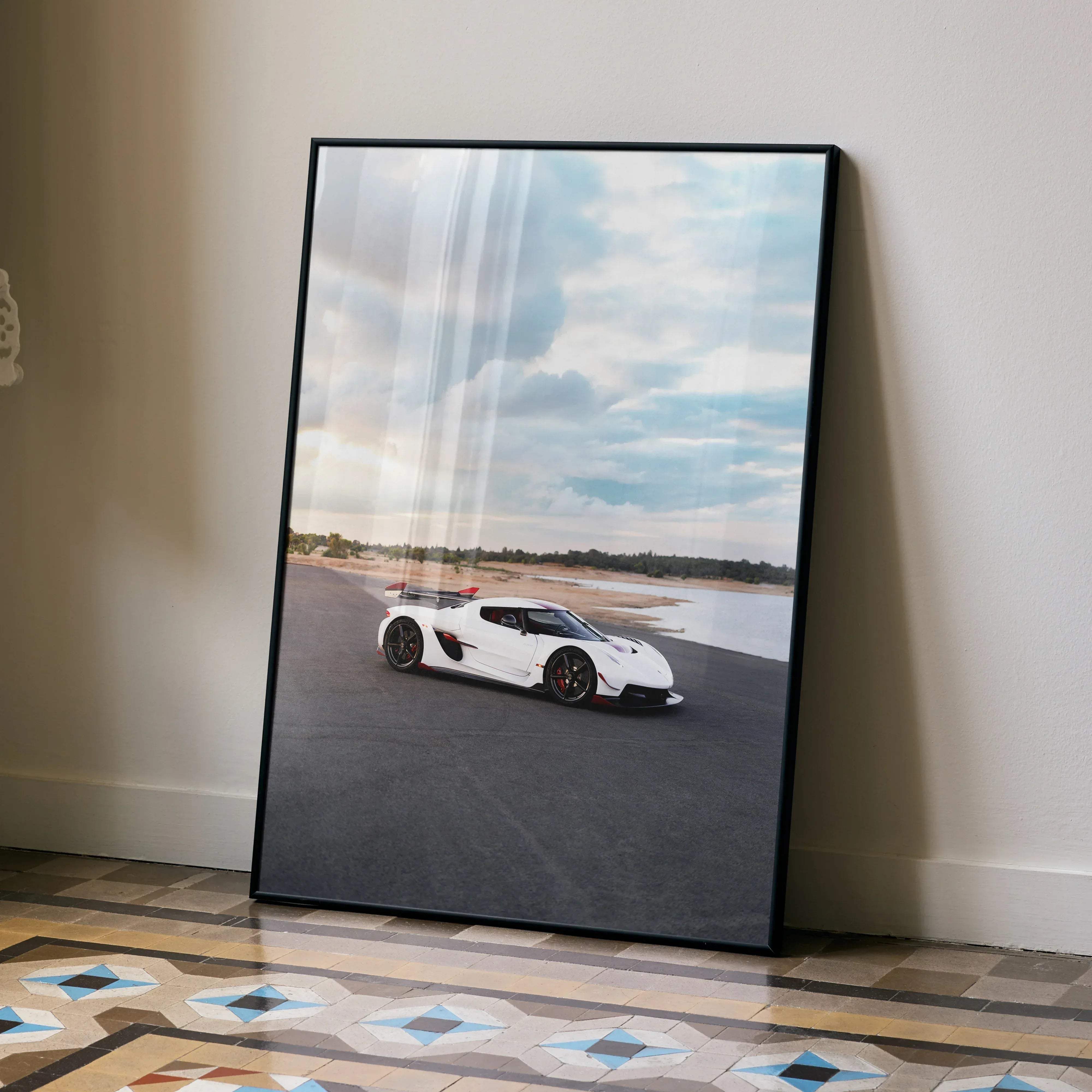 A framed poster of a white koenigsegg jesko hypercar with red accents on an asphalt track under a cloudy sky