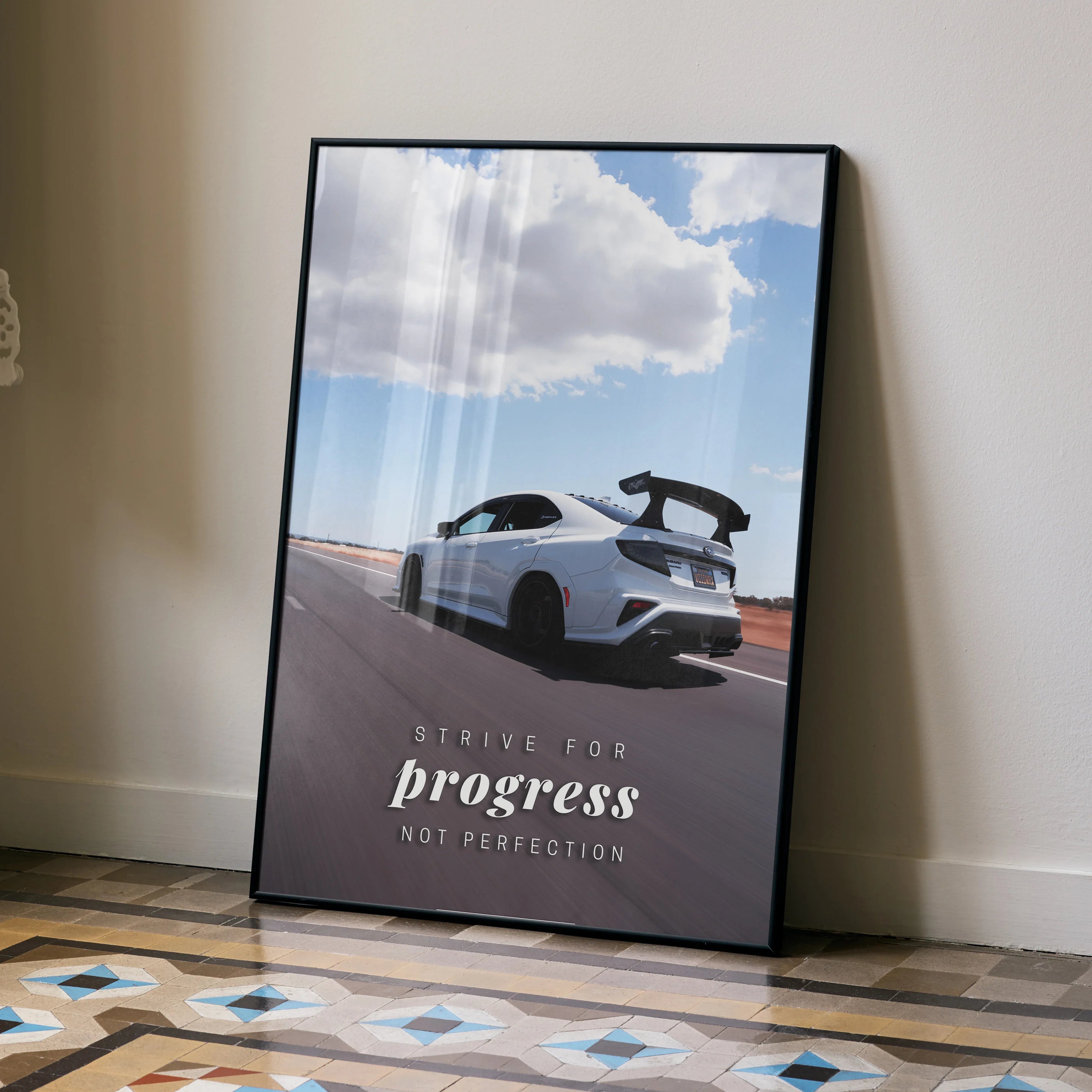 Poster wall art featuring a white Subaru WRX STI on a road under a blue sky with clouds and the quote "Strive for progress.