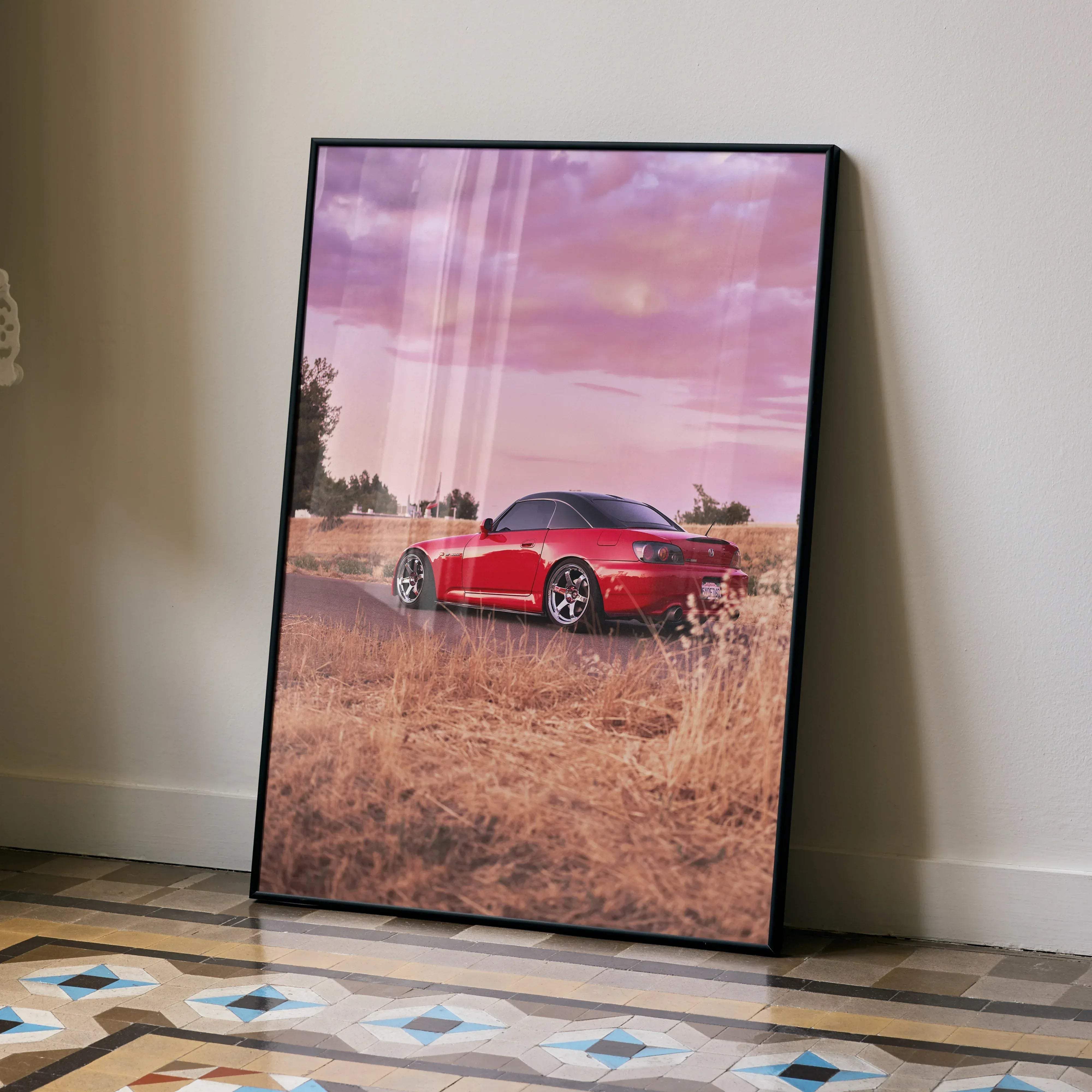 A framed poster of a red Honda S2000 sports car with black roof and silver rims against a pink sky over dry grass