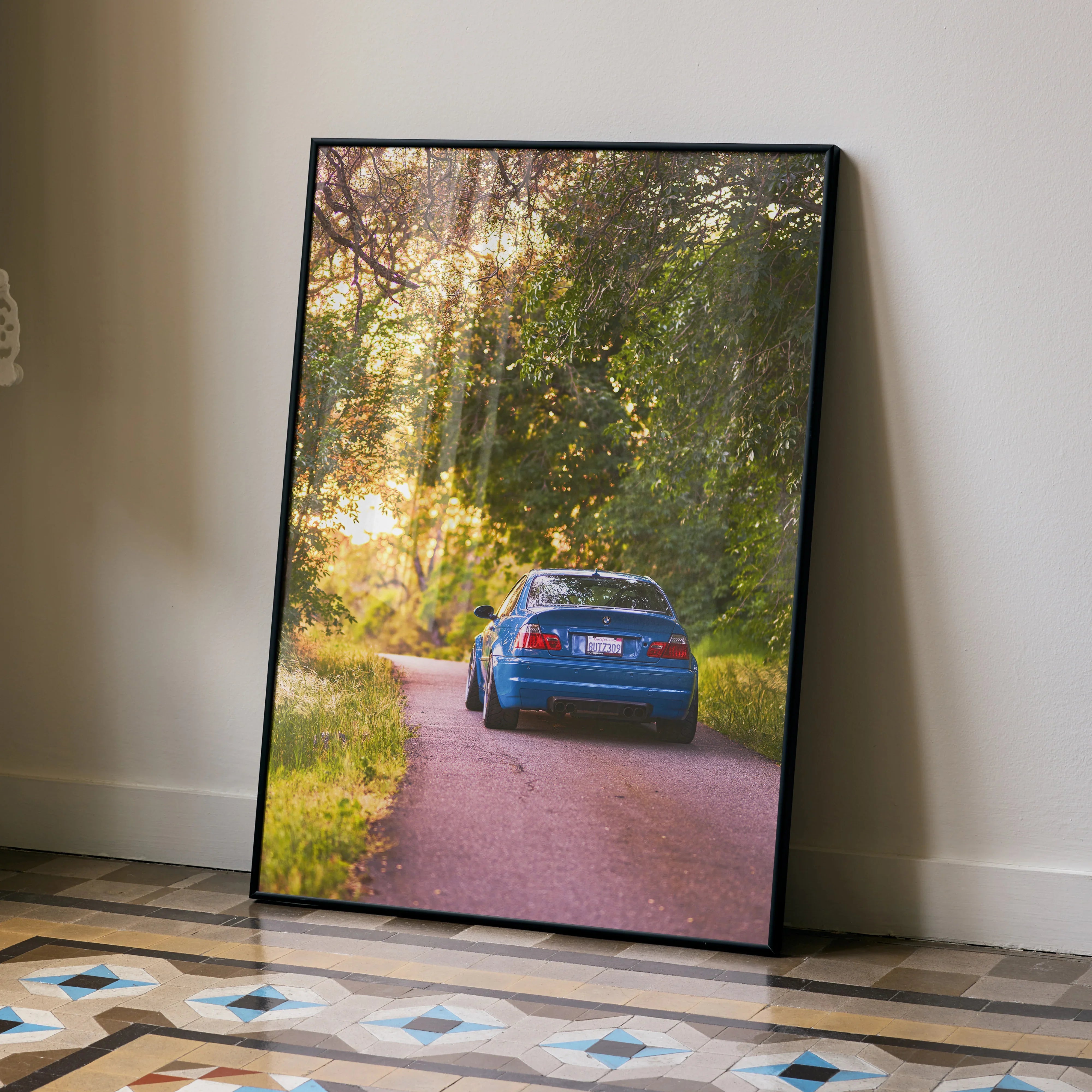 BMW E46 M3 on forest road at sunset poster wall art in black frame leaning on wall.