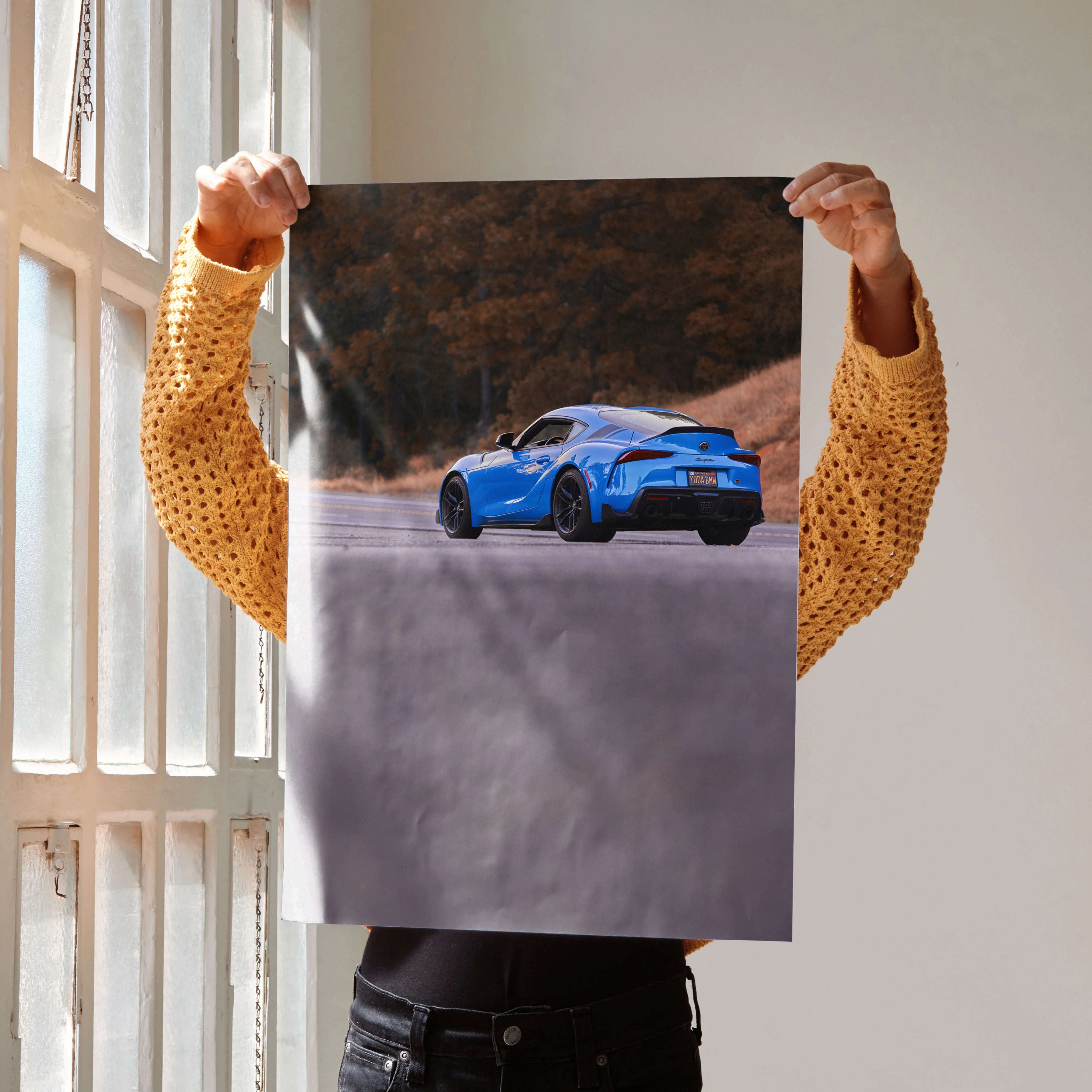 Blue Toyota Supra MK5 driving on scenic road poster wall art held against bright window light.
