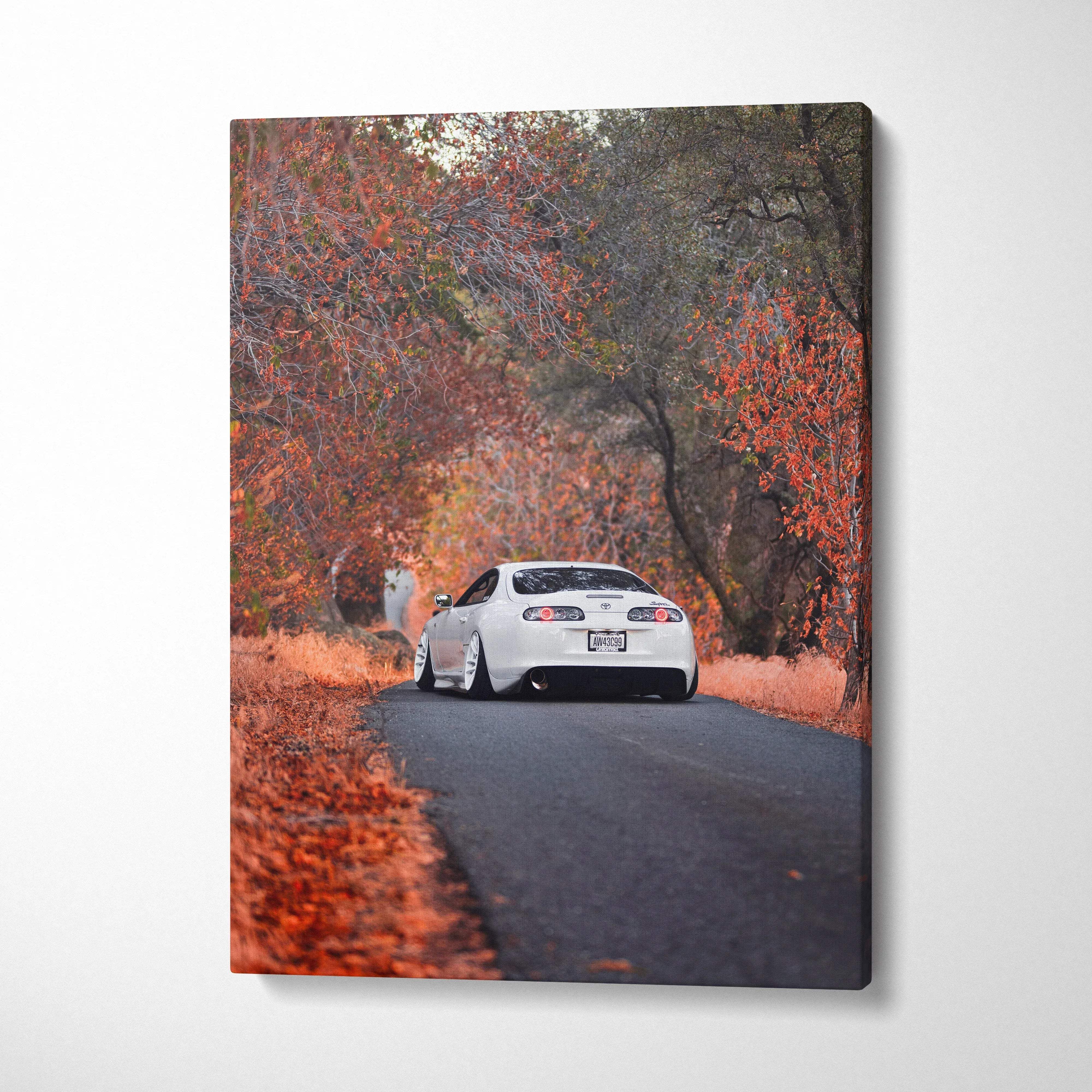 Vertical canvas wall art featuring a white Toyota Supra MK4 driving on a road surrounded by autumn trees with red foliage.