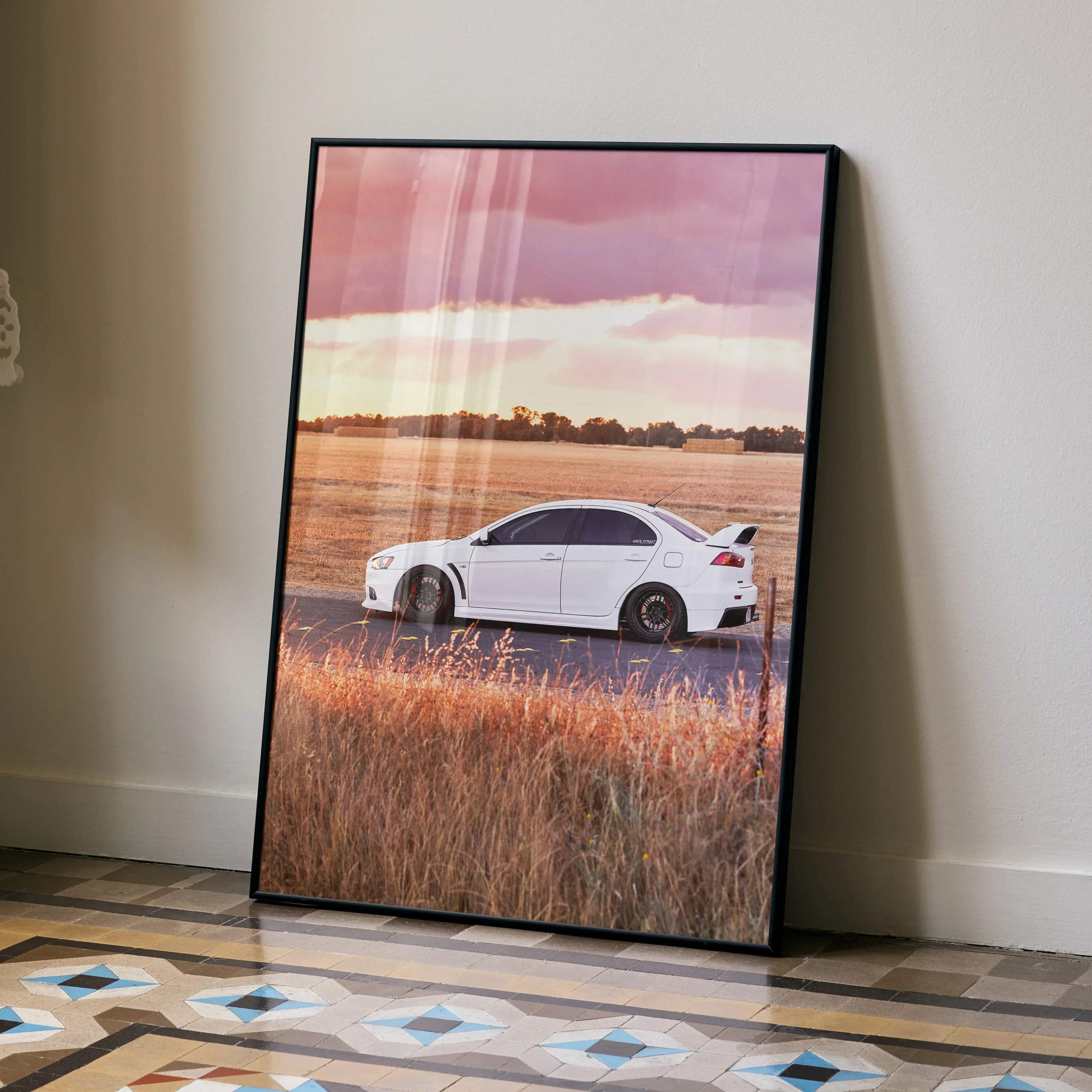 A framed Mitsubishi Evo 10 high resolution car poster showing a white sports car on a road with golden grass and a colorful sky