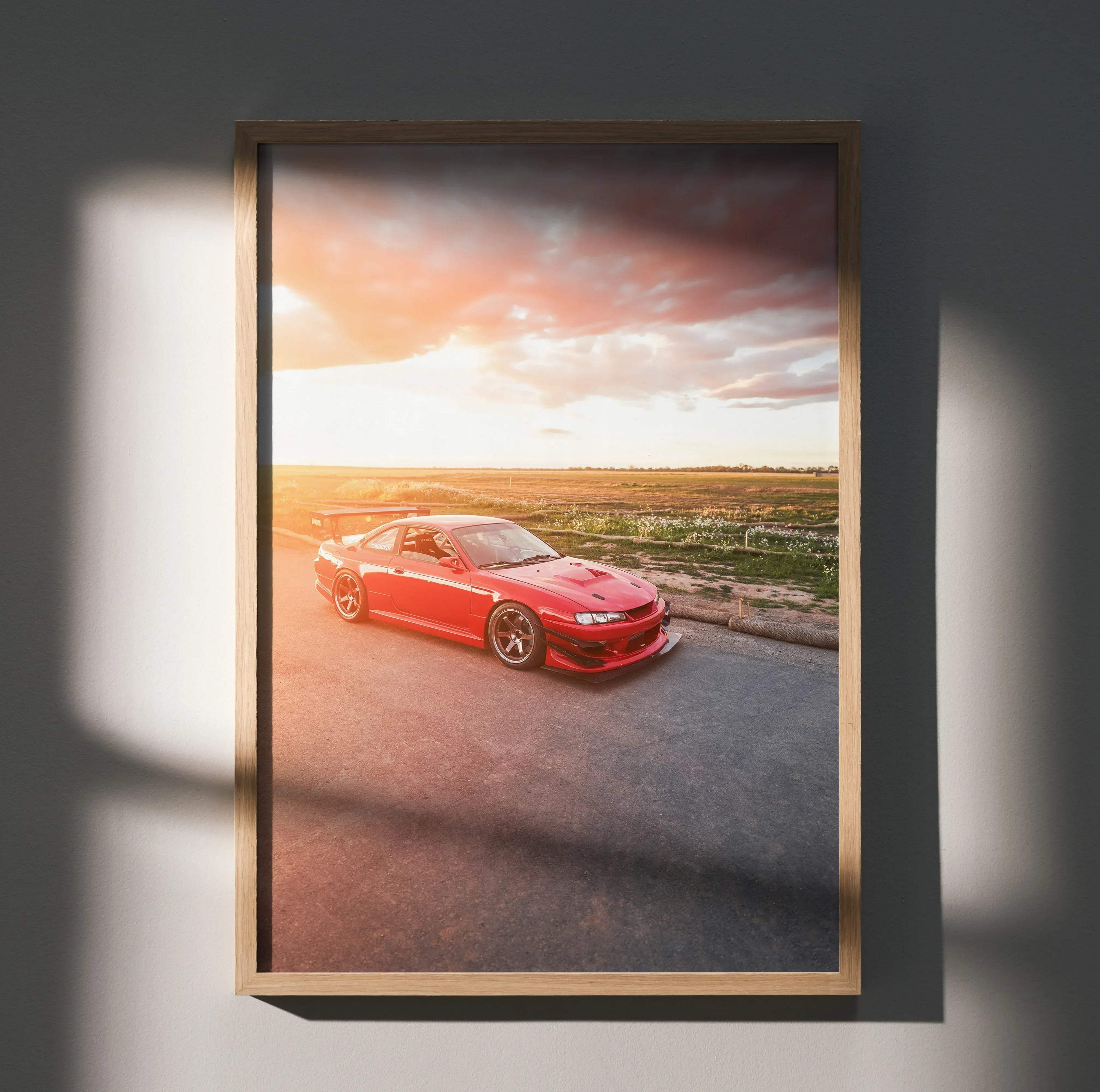A framed light wood poster of a red Nissan 240sx S14 Kouki sports car on a road at sunset