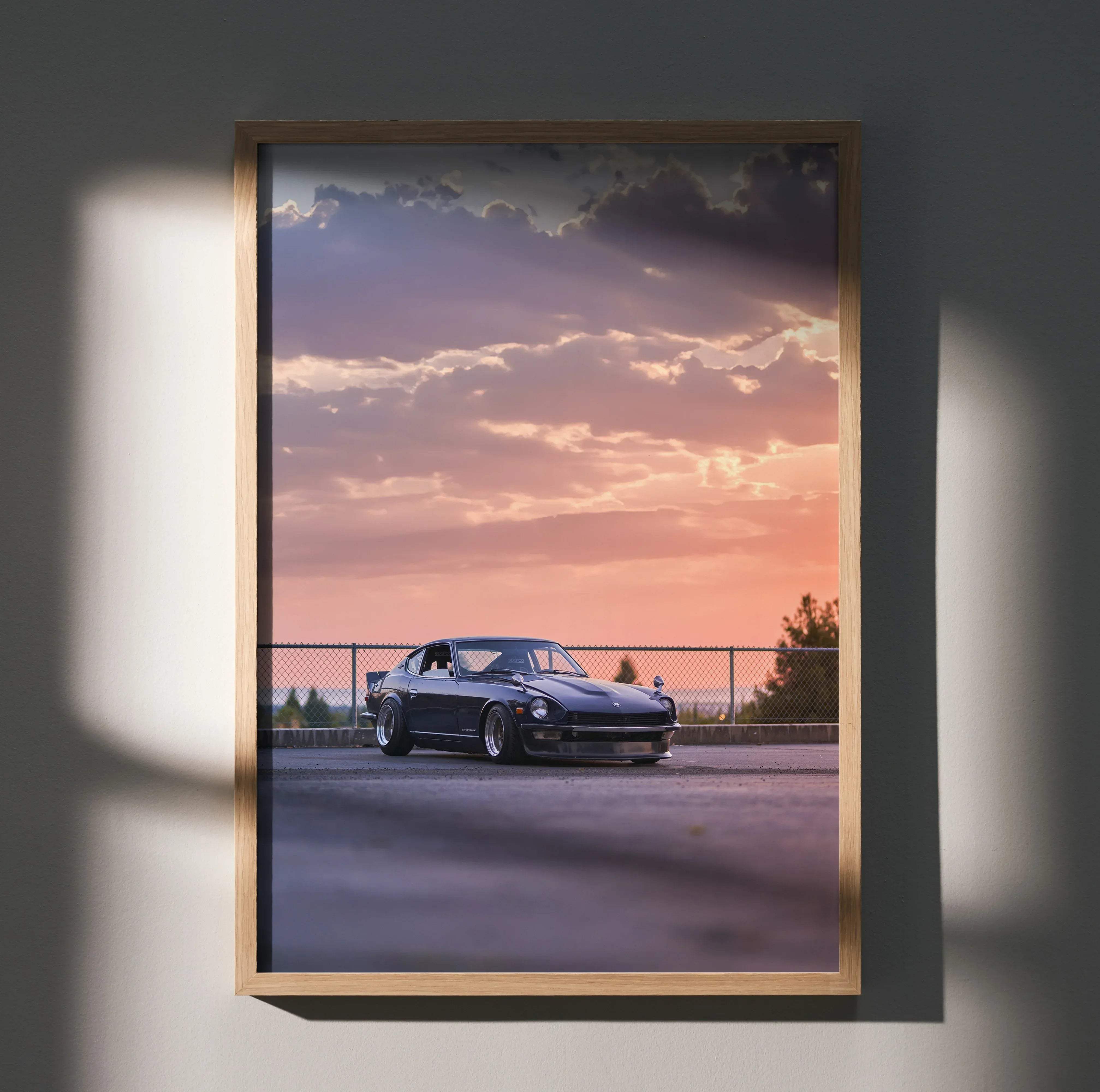 A framed poster of a dark blue Nissan 240z sports car under a vibrant pink and purple sunset sky