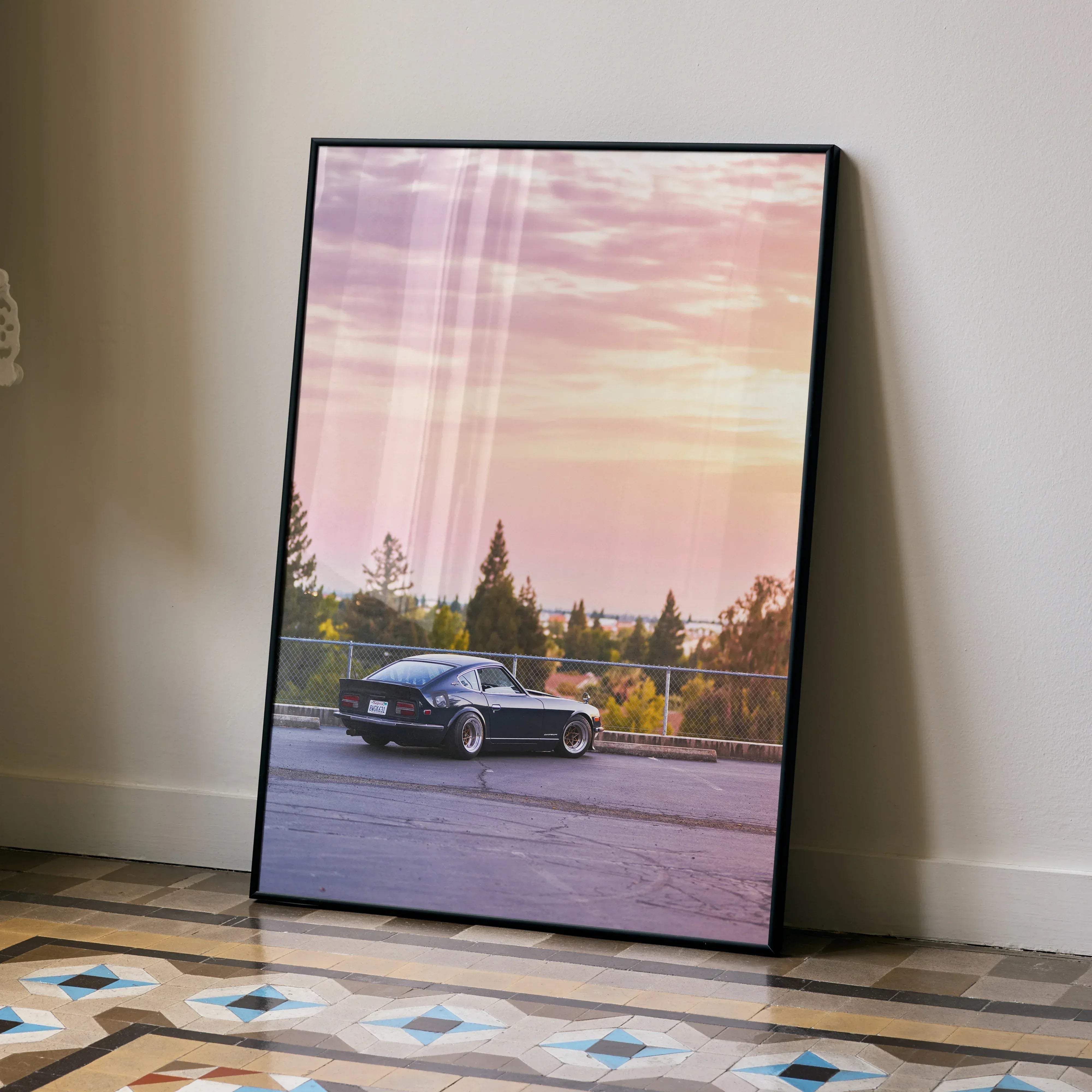 Black framed art print of a classic black Nissan 240z car on asphalt road under pink and orange sunset sky