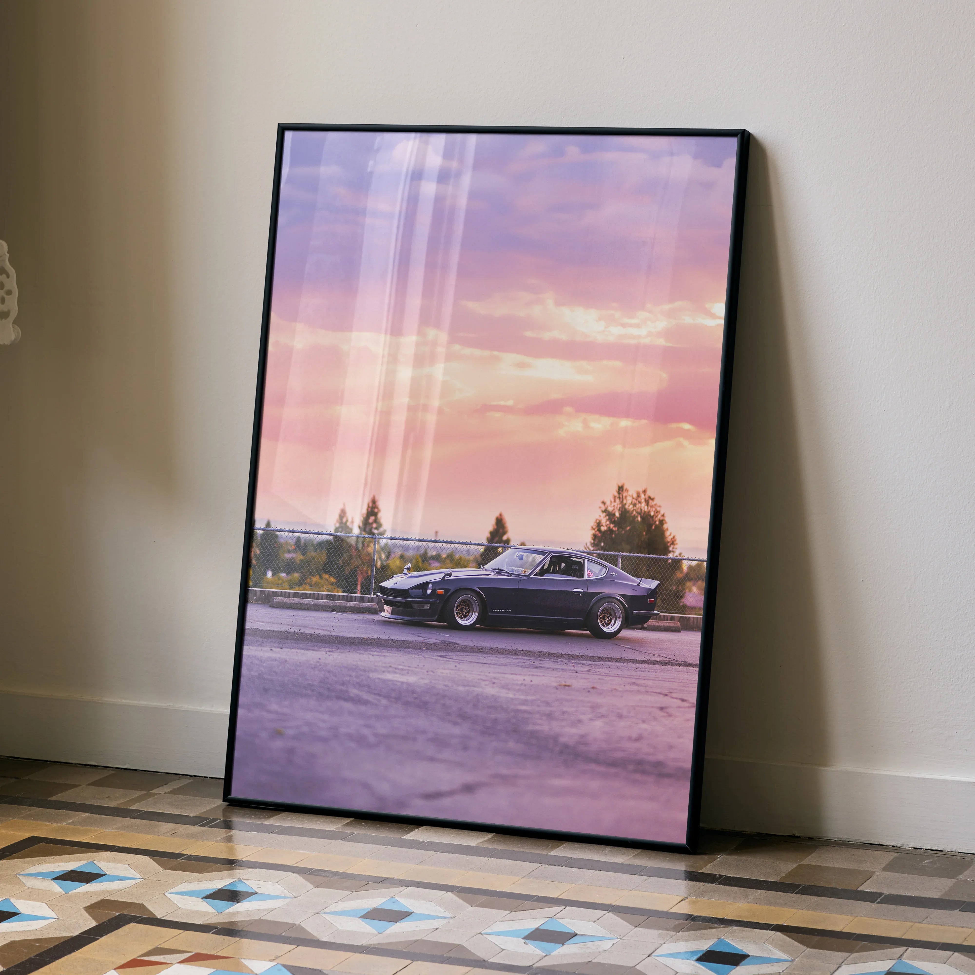 Nissan 240z poster wall art with vintage car and sunset sky in black frame resting on floor.