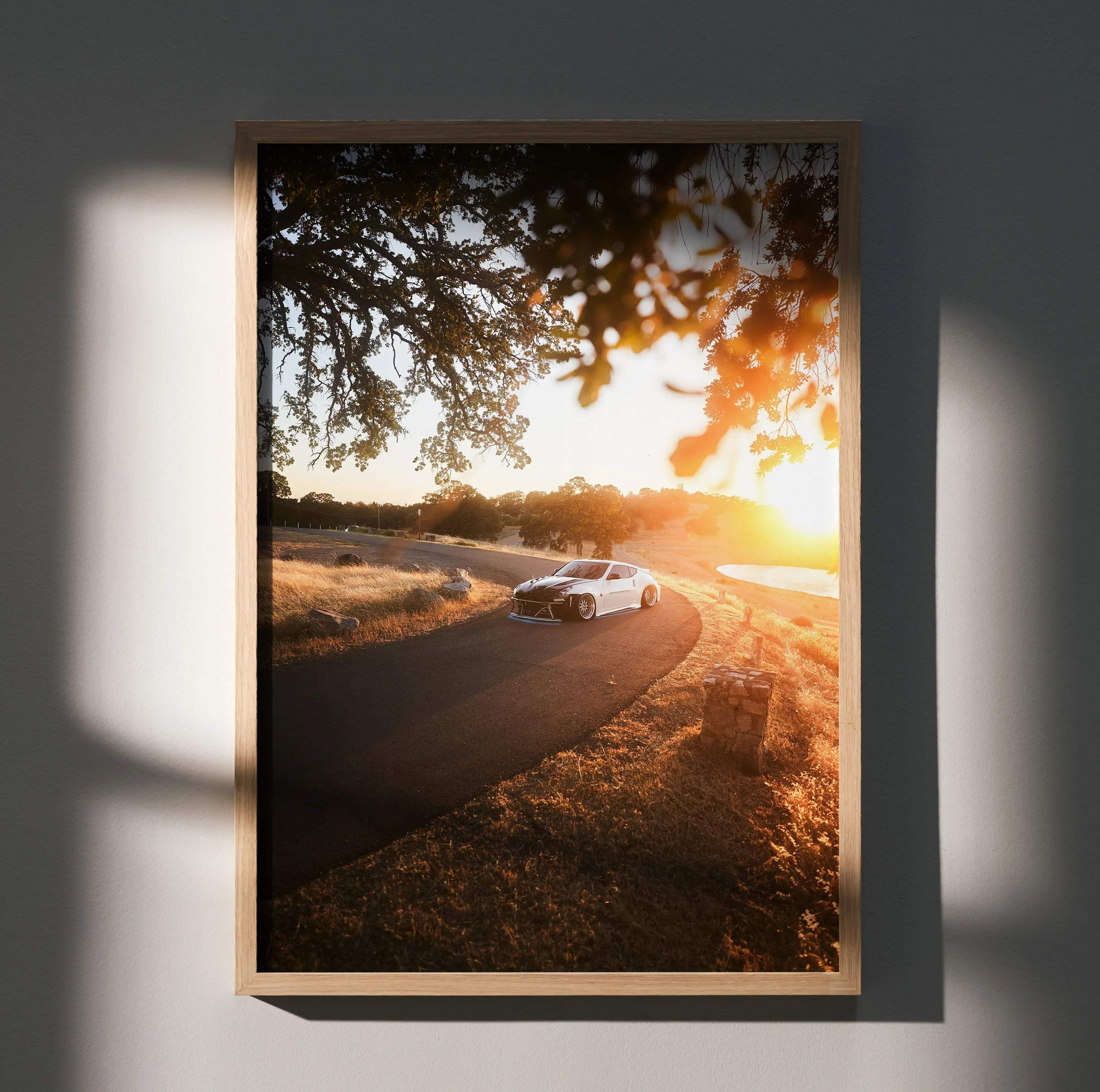 A framed photograph of a white Nissan 370z sports car on a winding road at sunset in a light wood frame