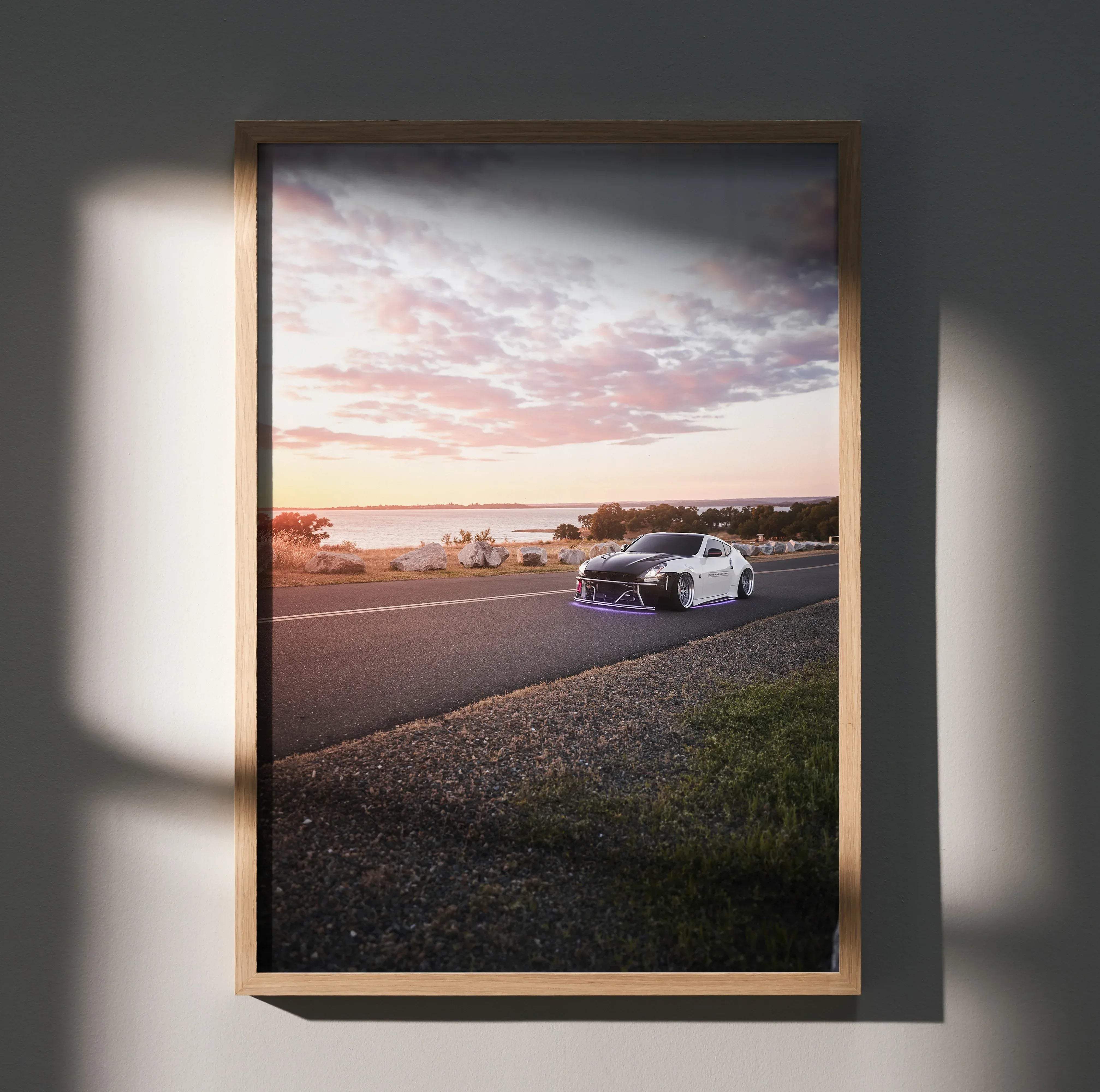 A framed high resolution car poster of a white and black Nissan 370z with purple underglow on a coastal road at sunset