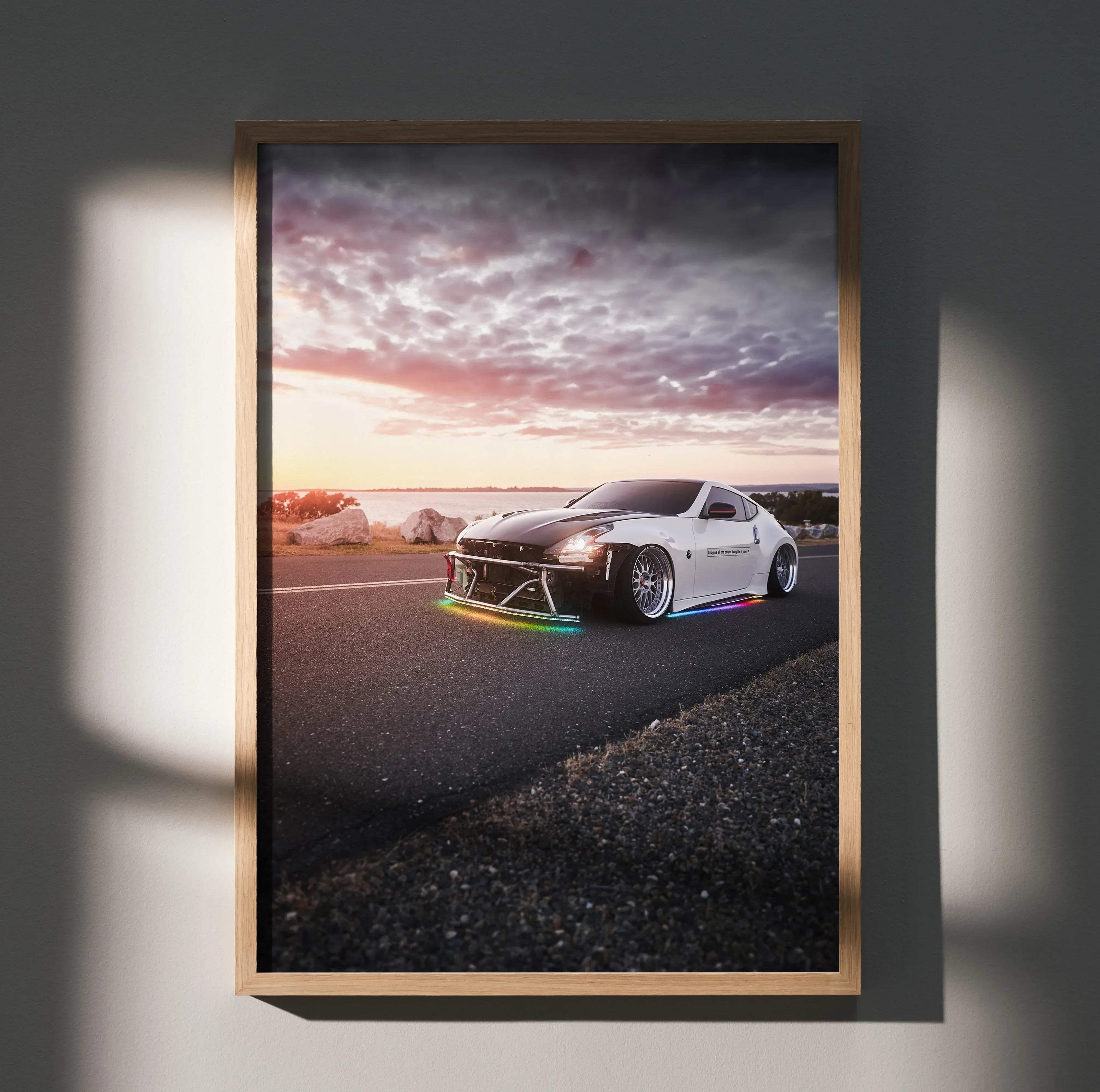 A framed poster of a white and black nissan 370z with rainbow led underglow on a coastal road at sunset