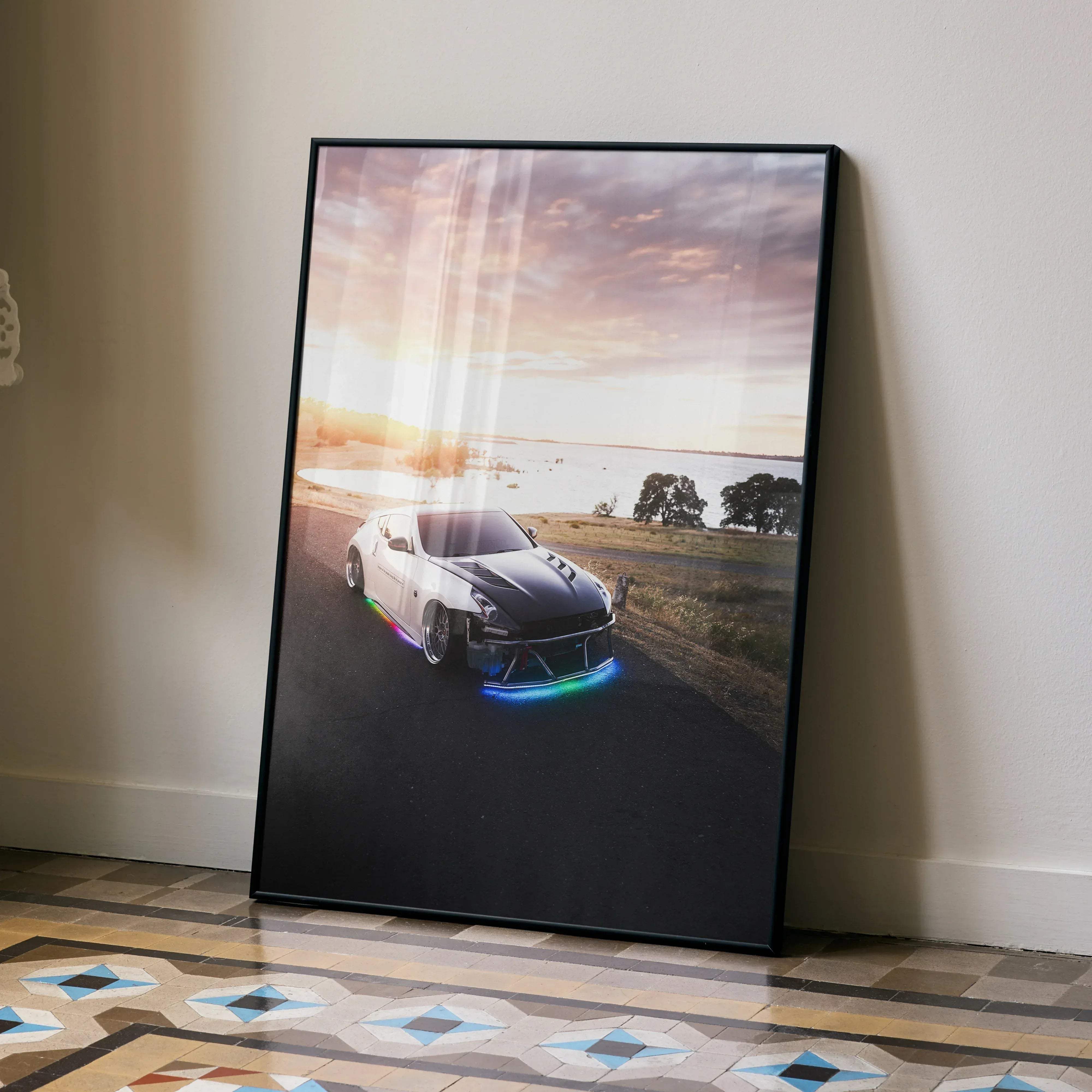 Black-framed poster of a white and black nissan 370z with glowing rainbow led lights on a sunset coastal road