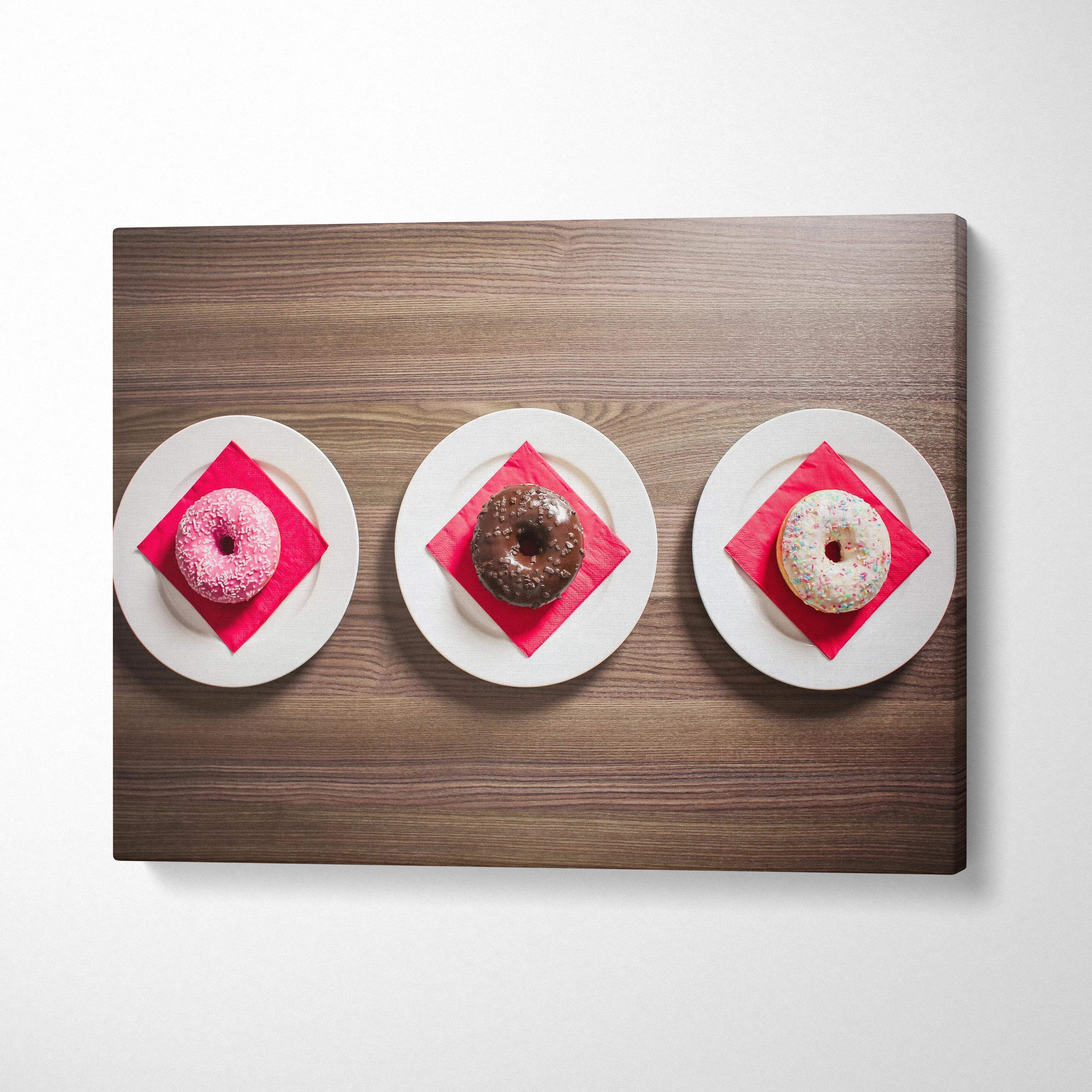 Canvas wall art featuring three vibrant glazed donuts on white plates with red napkins on wood table decor.
