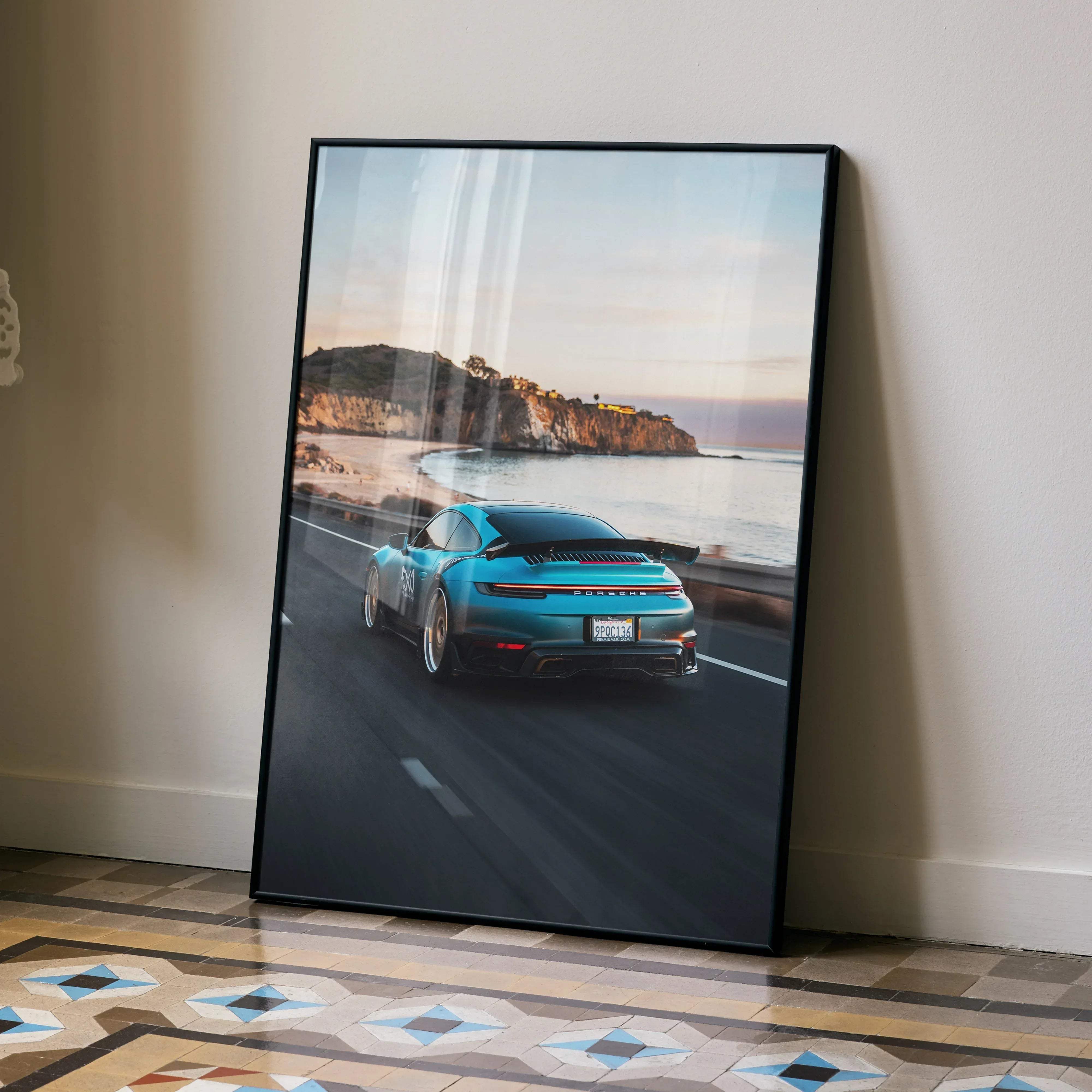 A framed poster of a turquoise Porsche 911 speeding on a coastal road at sunset in a sleek black frame