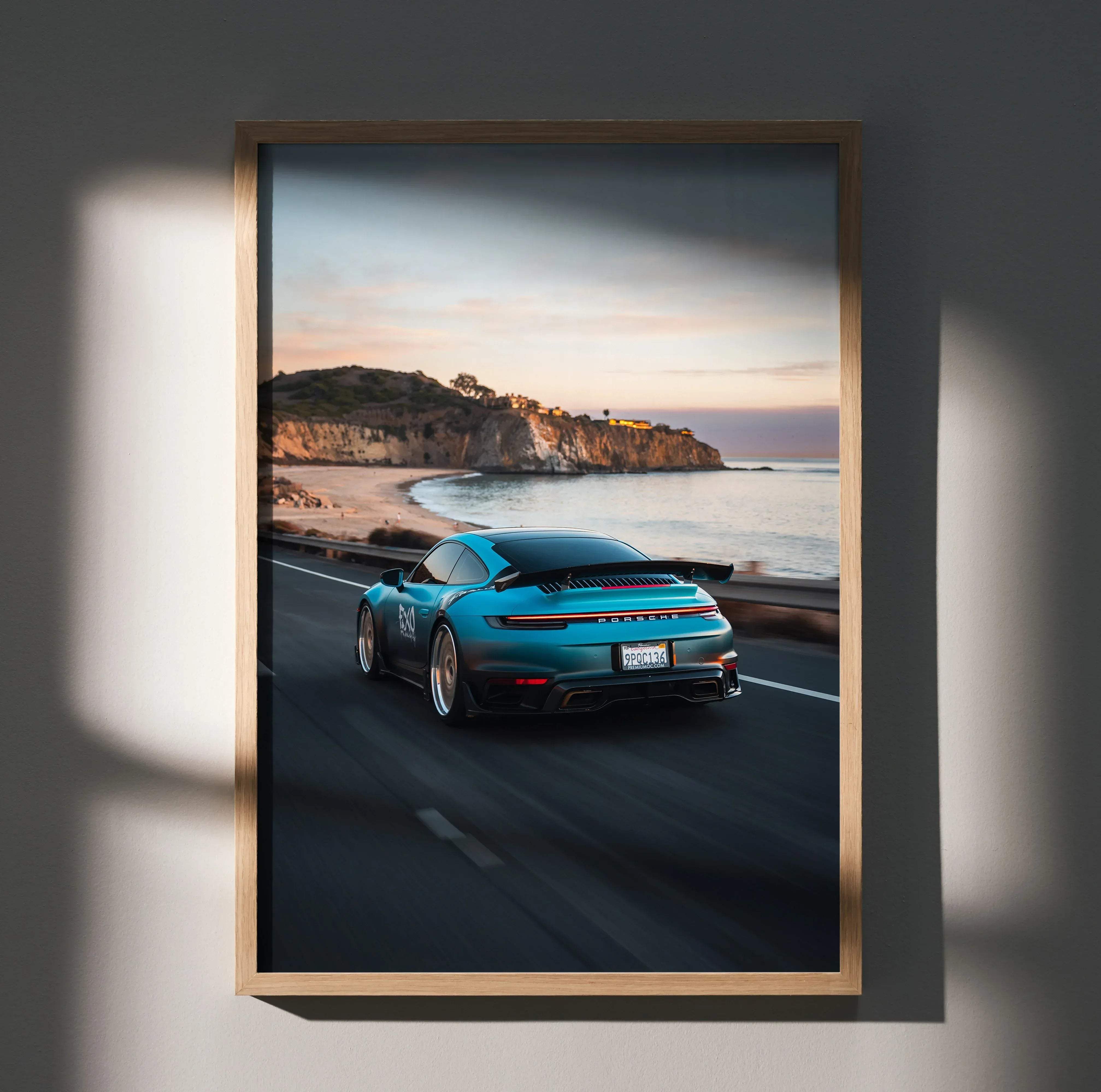 A framed car poster featuring a turquoise Porsche 911 speeding along a coastal road at sunset
