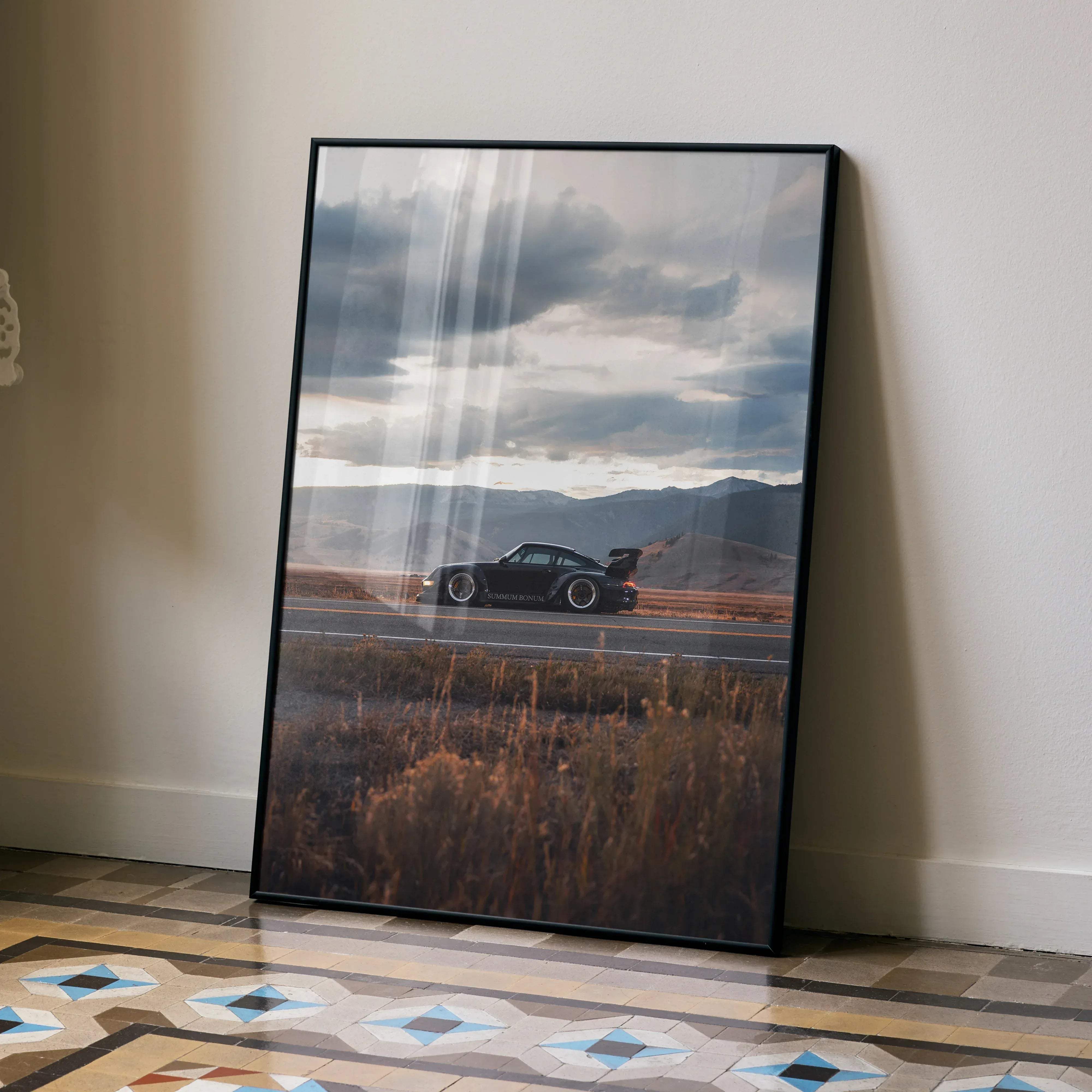 Black framed art print of a sleek black porsche 911 speeding on a winding road under dramatic cloudy skies
