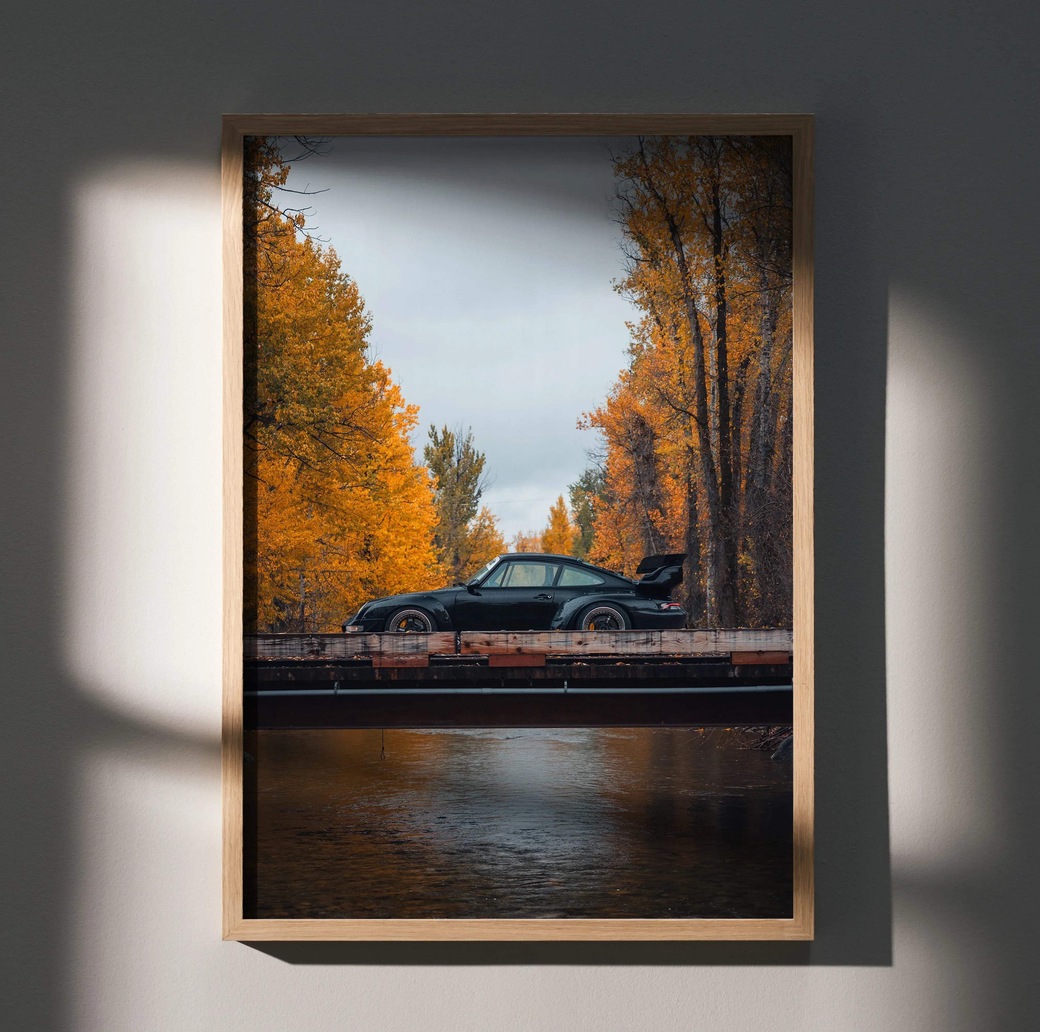 A framed photograph of a black Porsche 911 sports car with rear spoiler up on wooden bridge autumn trees backdrop