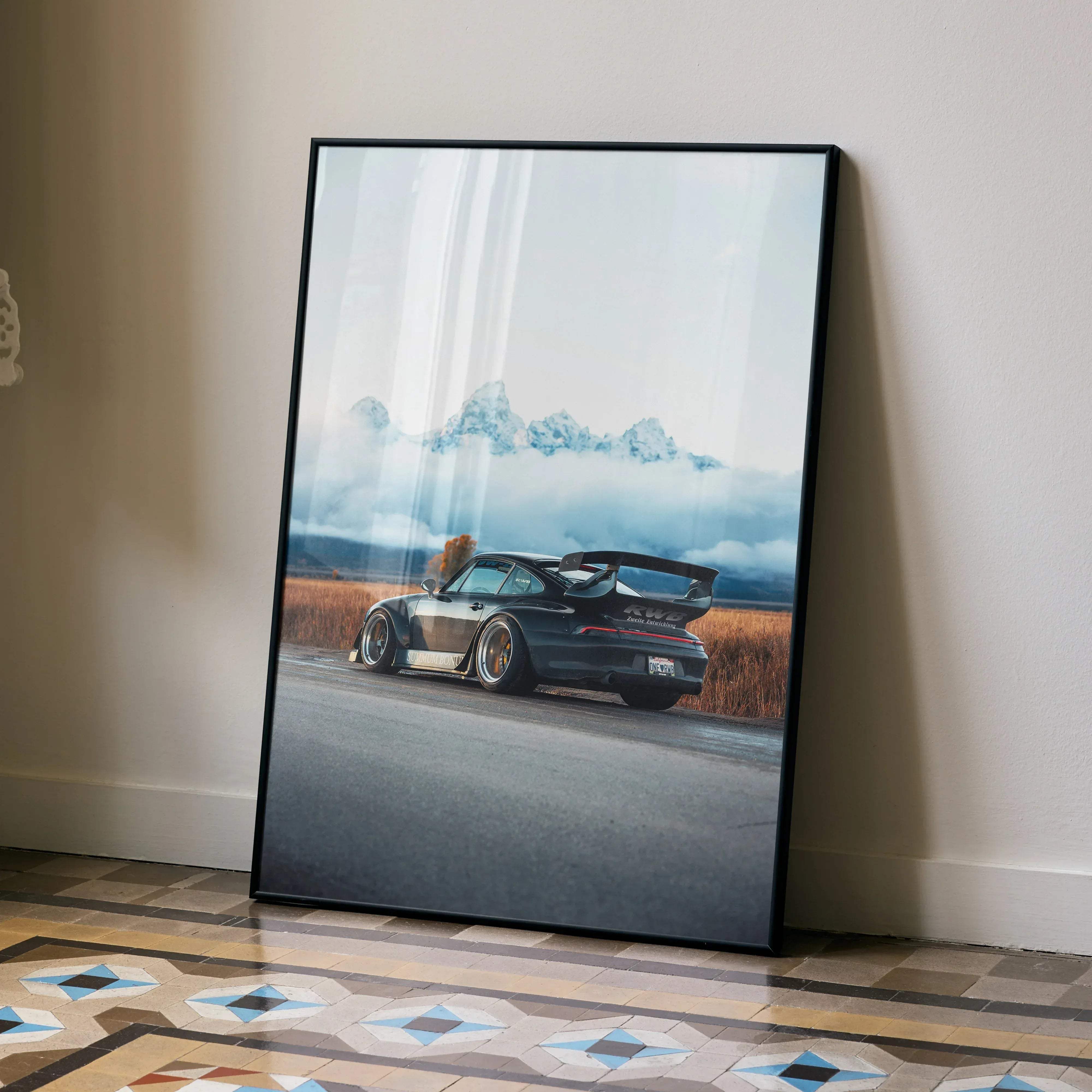 Black-framed poster of matte black rwb porsche 911 with gold rims and rear spoiler against misty mountains and grass