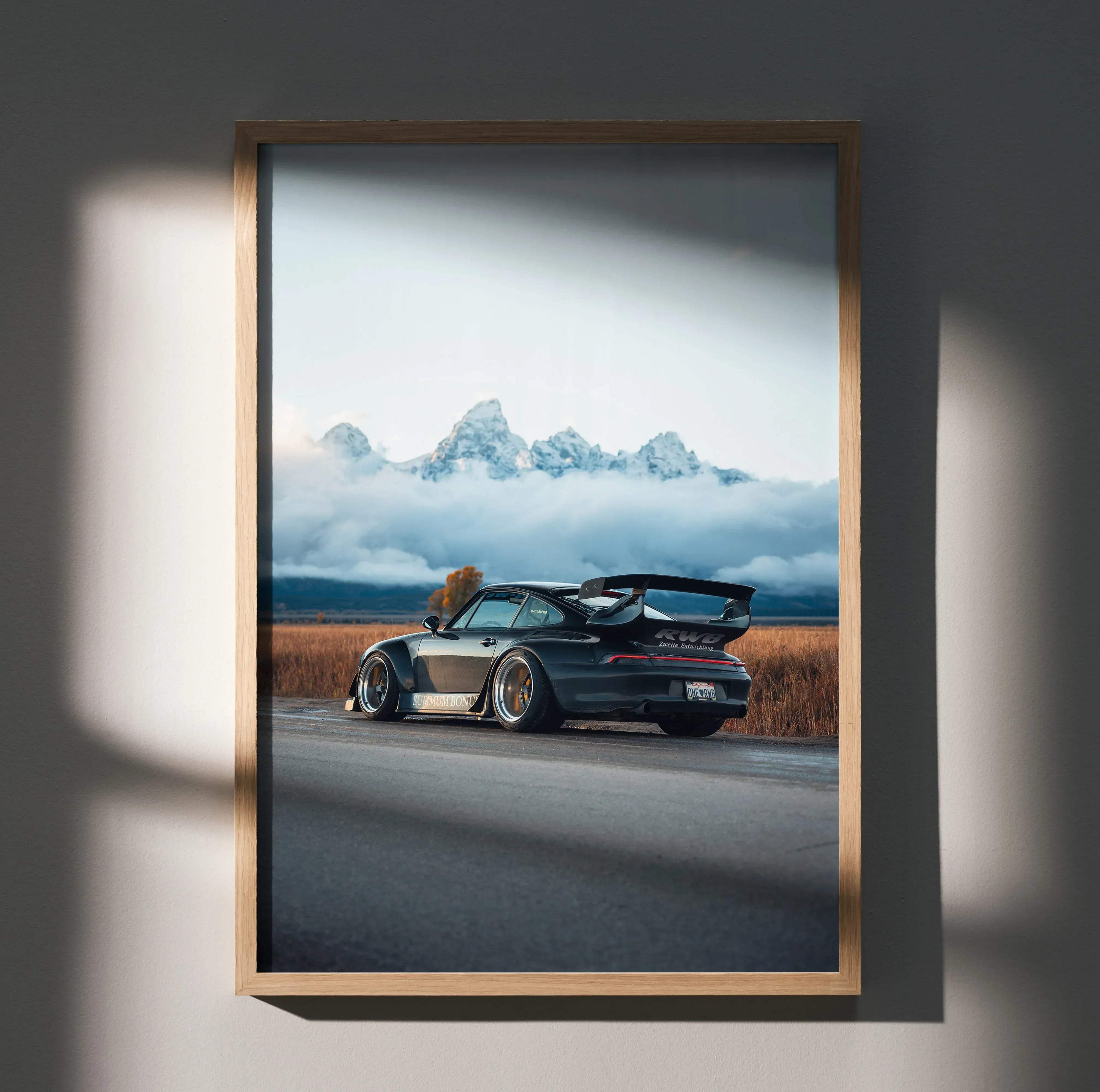 A framed photograph of a black rwb porsche 911 with a rear spoiler against misty mountains and golden fields poster