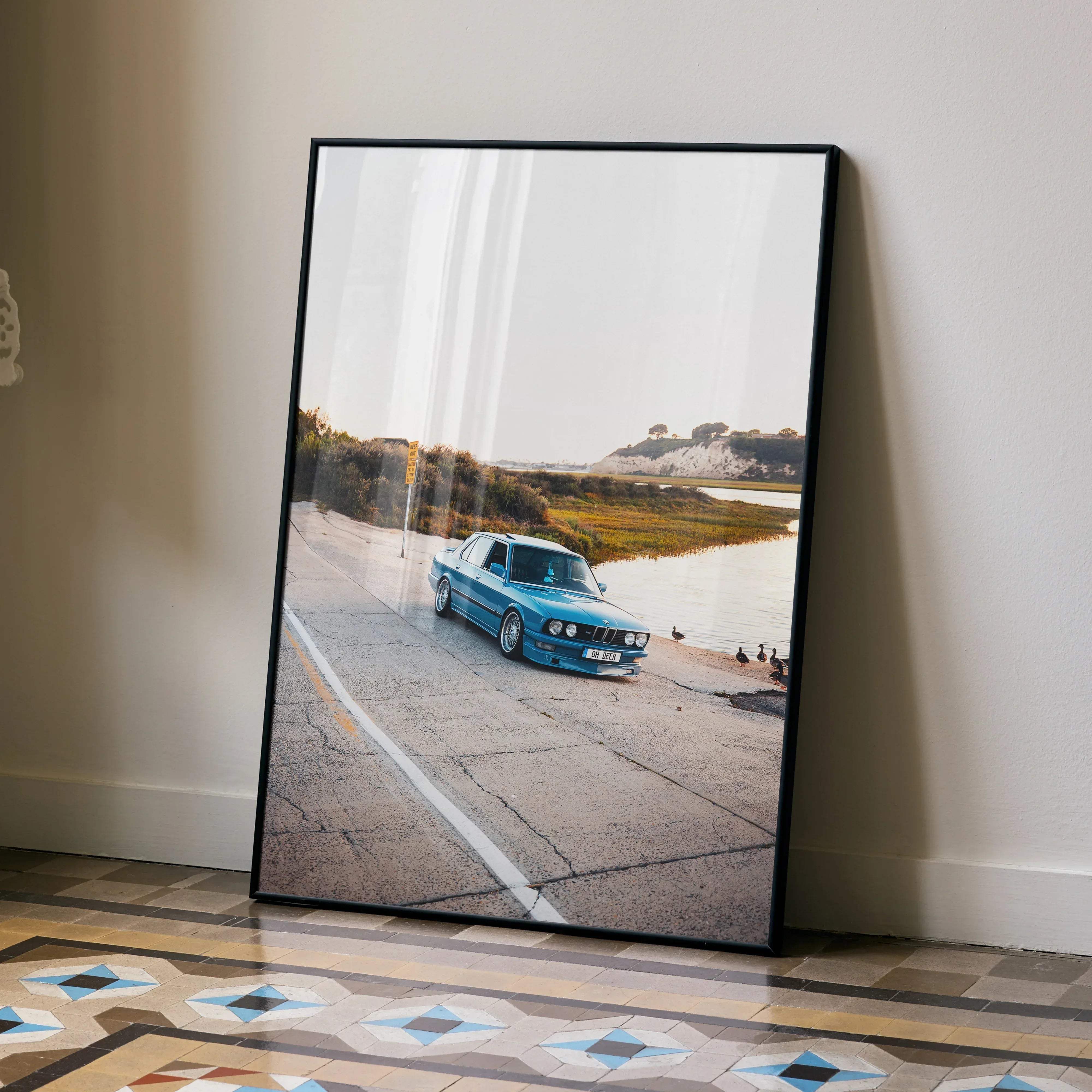 A framed high resolution art print poster of a vibrant blue BMW e30 sedan on a coastal road